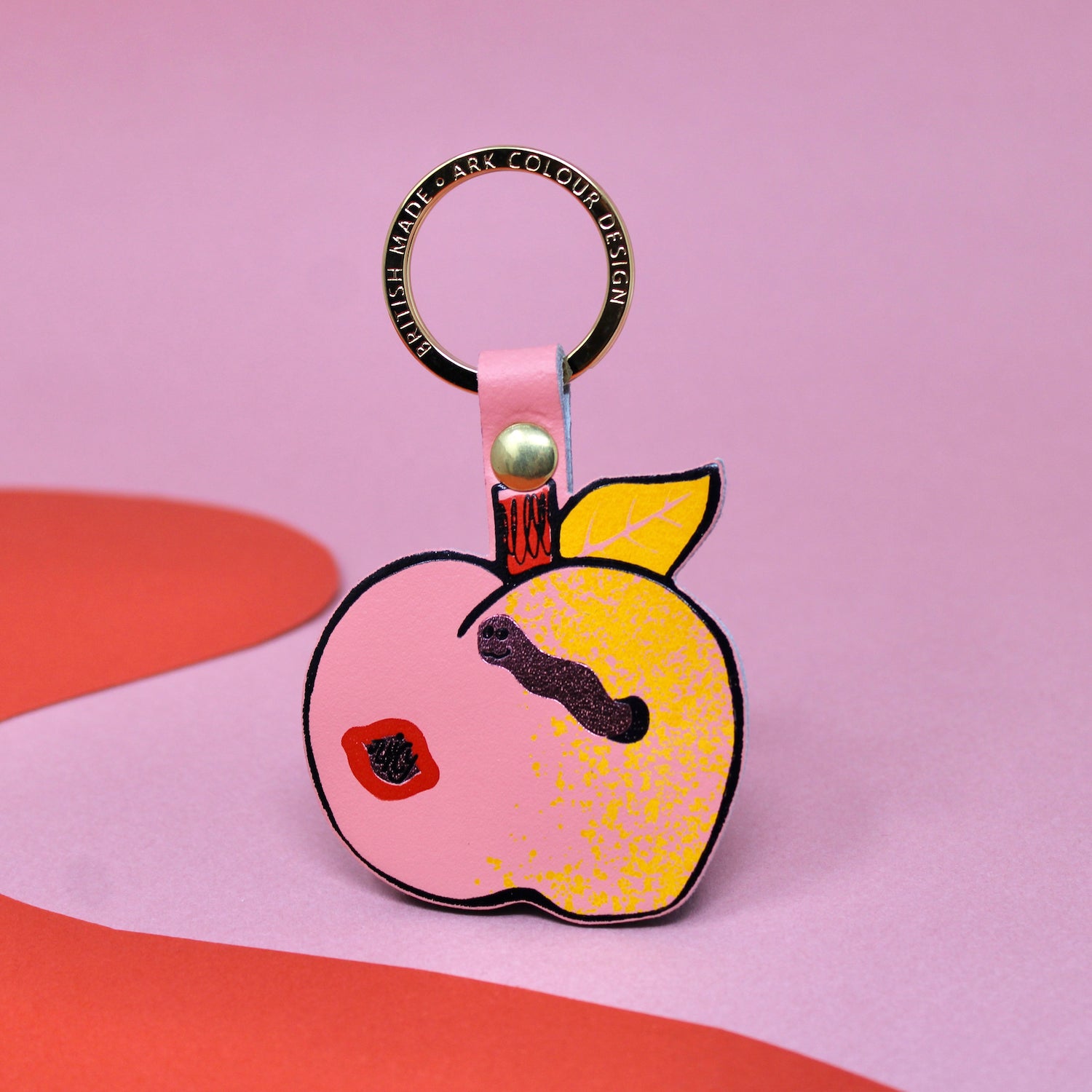 Ark Colour Design apple keychain with pink strap, gold ring, and illustrated red apple design on a pink and red background.