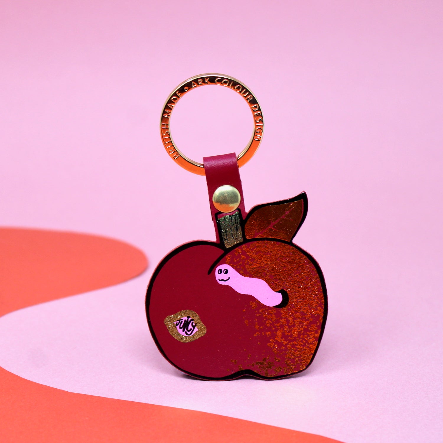 Ark Colour Design apple keychain with red strap, gold ring, and illustrated red apple design on a pink and red background.