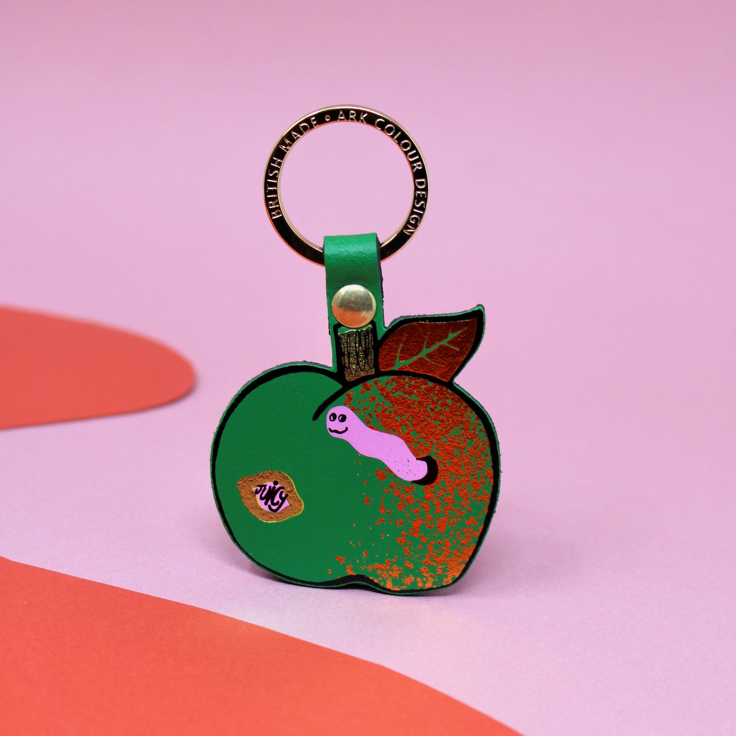 Ark Colour Design apple keychain with green strap, gold ring, and illustrated red apple design on a pink and red background.