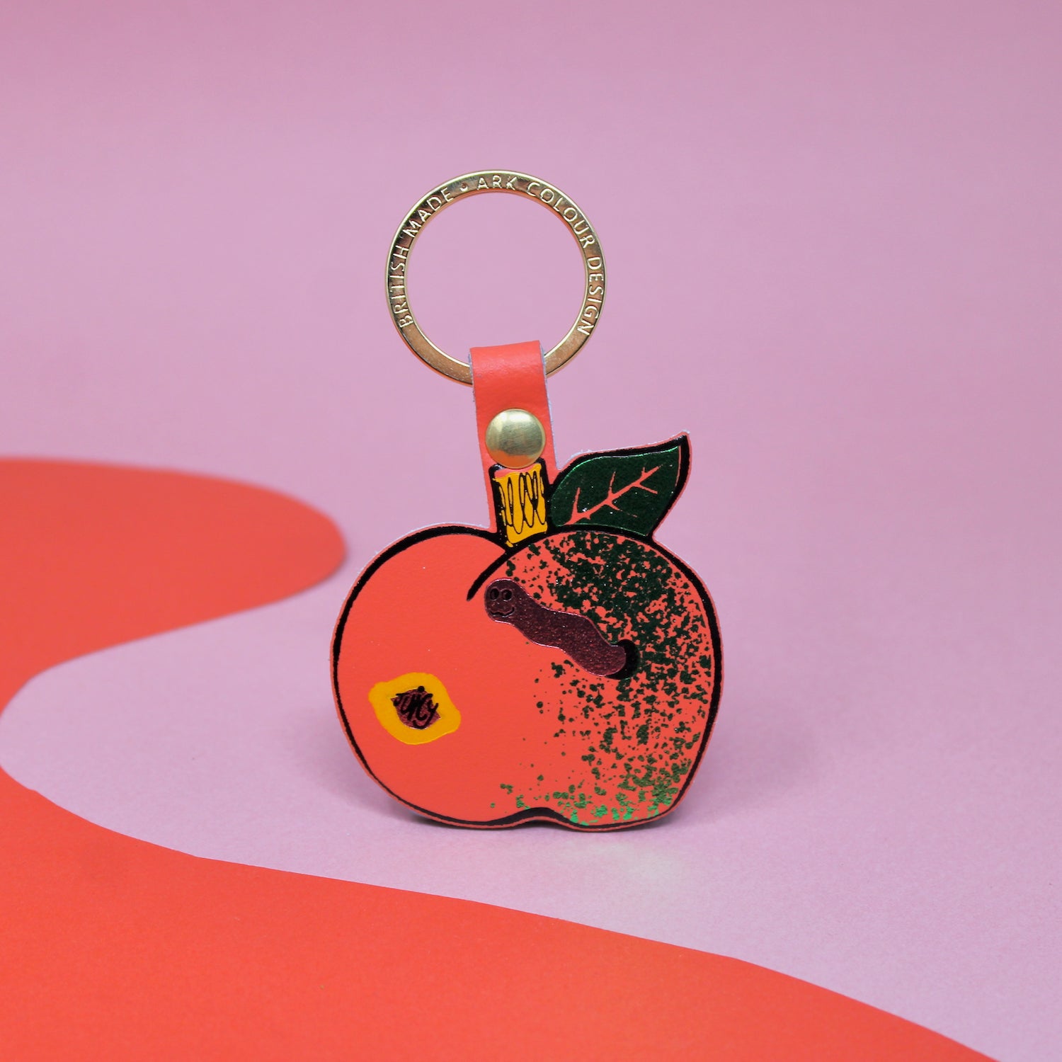 Ark Colour Design apple keychain with coral strap, gold ring, and illustrated red apple design on a pink and red background.