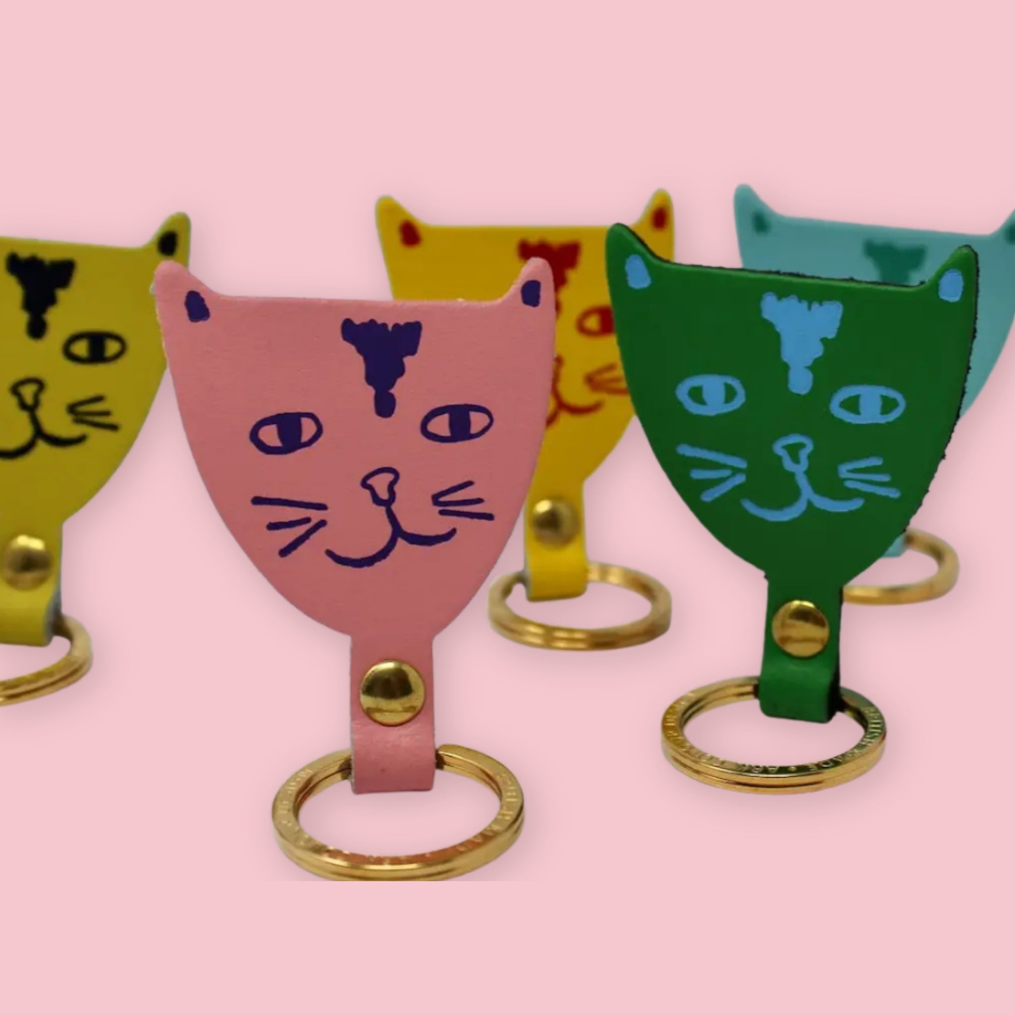 Ark Colour Design cat keychains in assorted colors, shown with gold rings and hand-drawn cat face designs on a pink background.