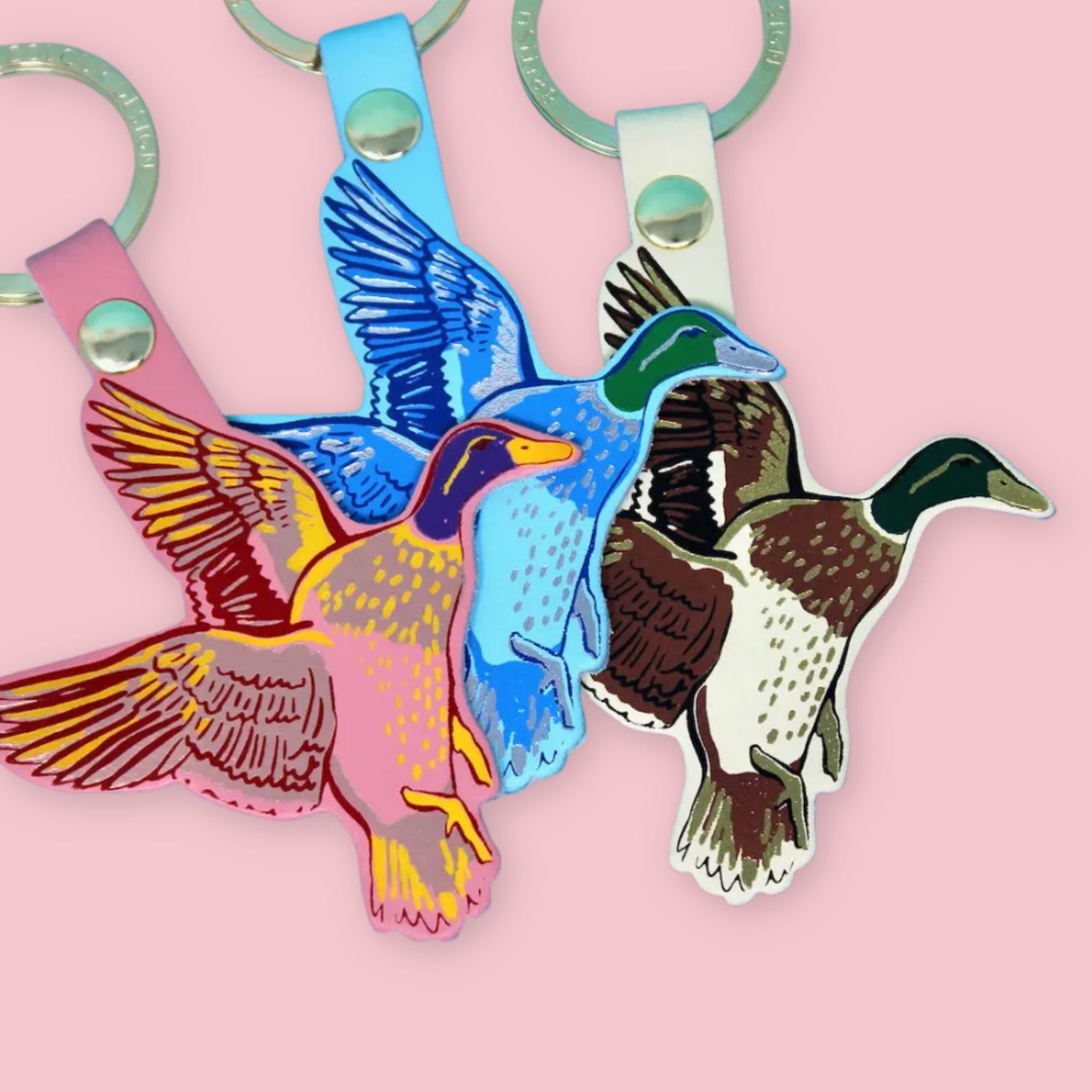 Ark Colour Design flying duck keychains in pink, blue, and cream colorways, shown with gold rings on a pink background.