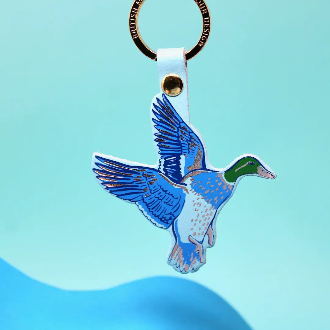Ark Colour Design flying duck keychain with turquoise strap, gold ring, and colorful bird illustration on a bright blue background.
