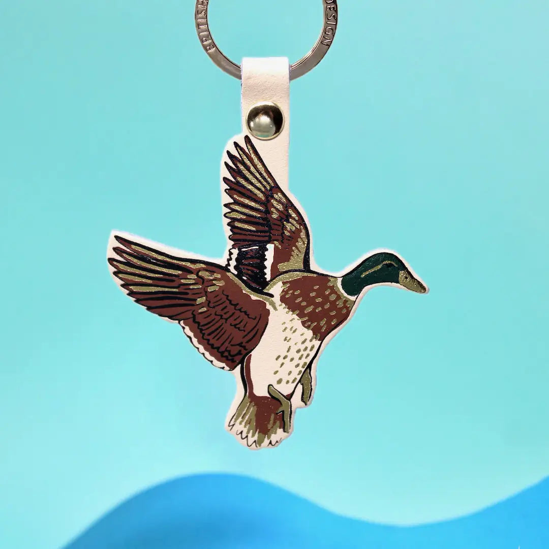 Ark Colour Design flying duck keychain with cream strap, gold ring, and colorful bird illustration on a bright blue background.