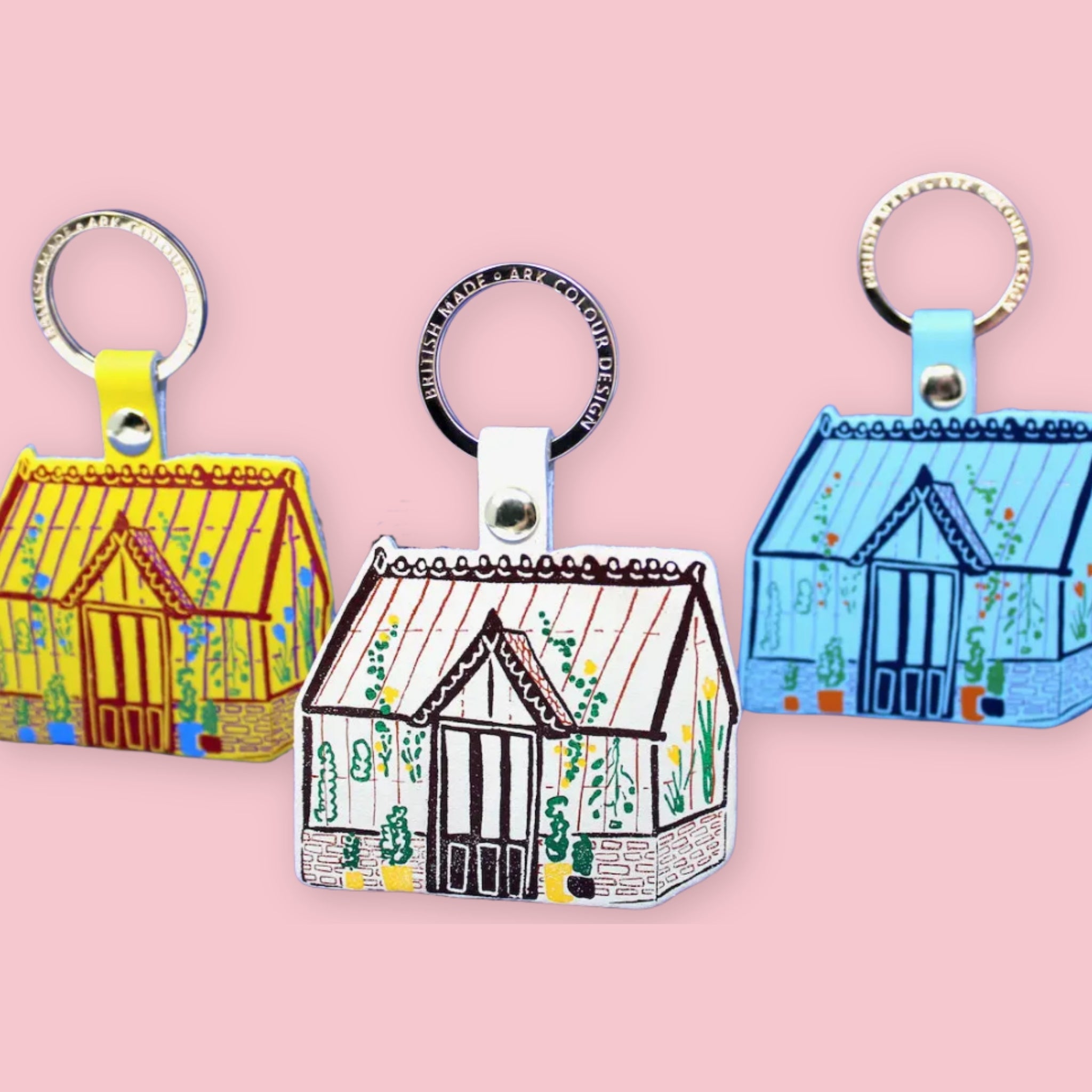 Ark Colour Design greenhouse keychains in yellow, white, and blue colorways, shown with gold key rings on a pink background.