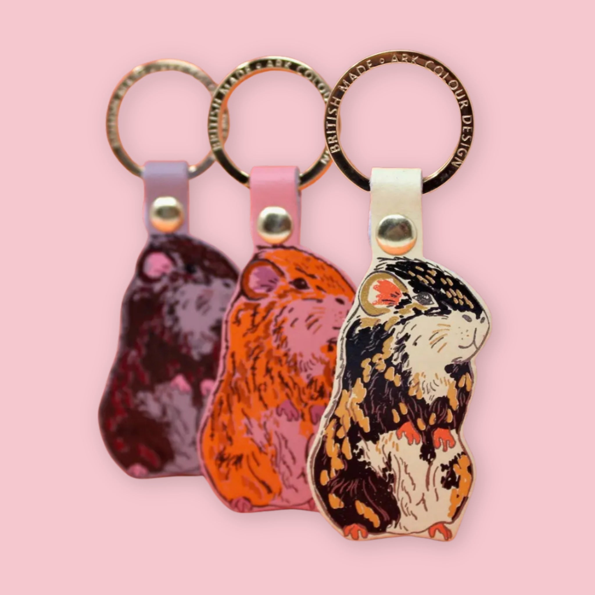 Ark Colour Design guinea pig keychains in three color styles, shown with gold key rings on a pink background.