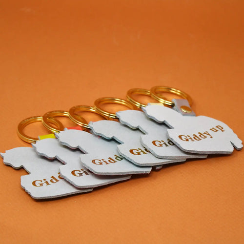 Back side of Ark Colour Design horse keychains with gold rings and “Giddy up” text on an orange background.