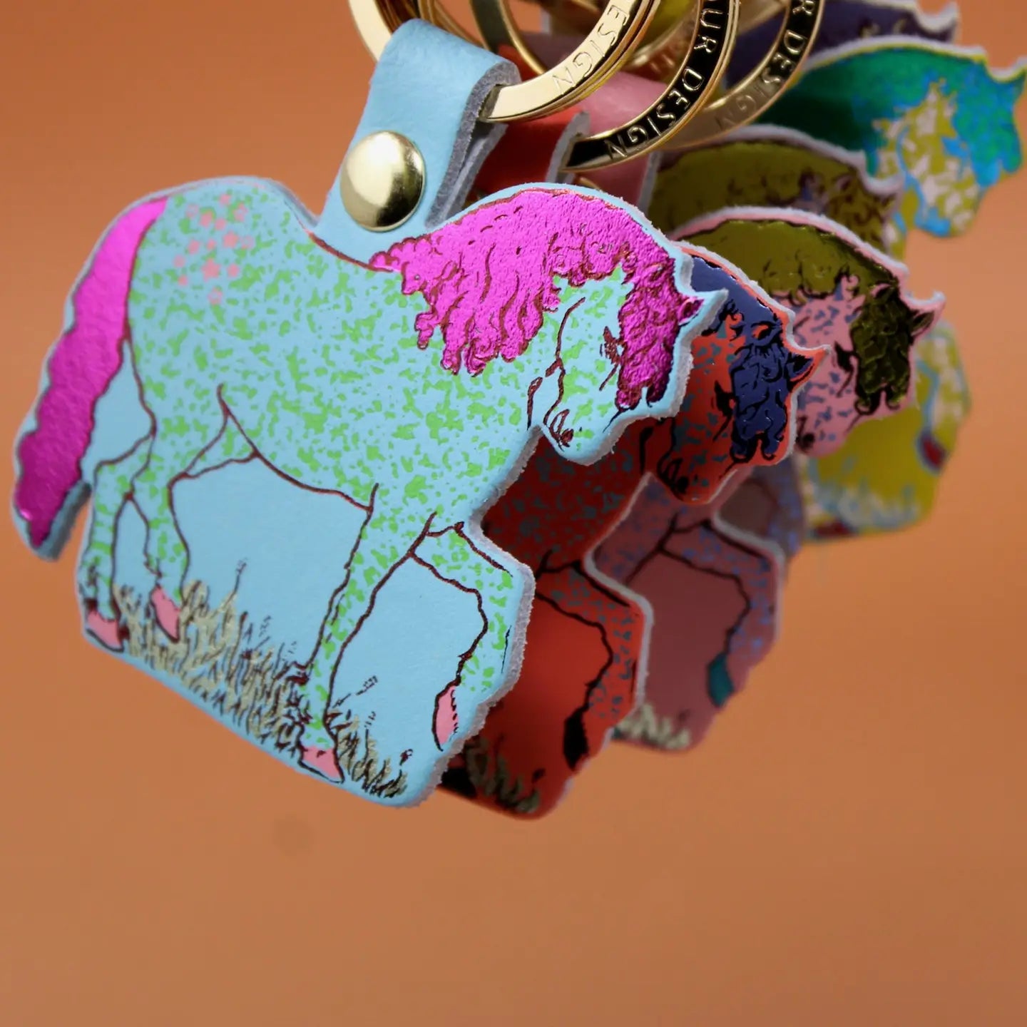 Close-up of colorful Ark Colour Design horse keychains with gold rings, shown layered on an orange background.