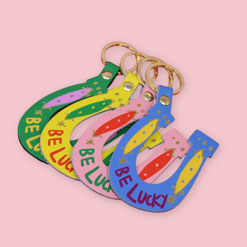 Ark Colour Design horseshoe keychains in green, yellow, pink, and blue, each with “Be Lucky” text and gold key rings.