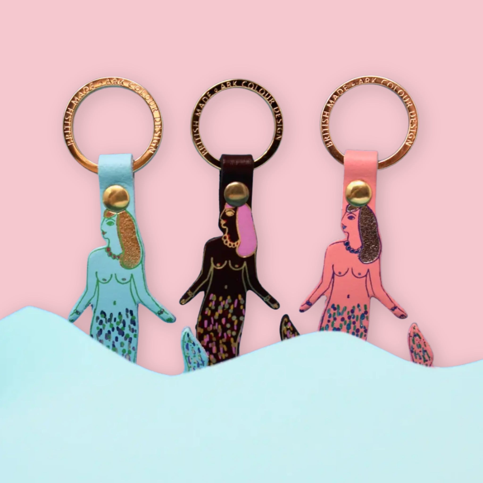 Three mermaid-themed keychains lined up against a pink background with a light blue wave shape at the bottom. The keychains have gold-tone rings and are shown in light blue, burgundy, and pink colorways.