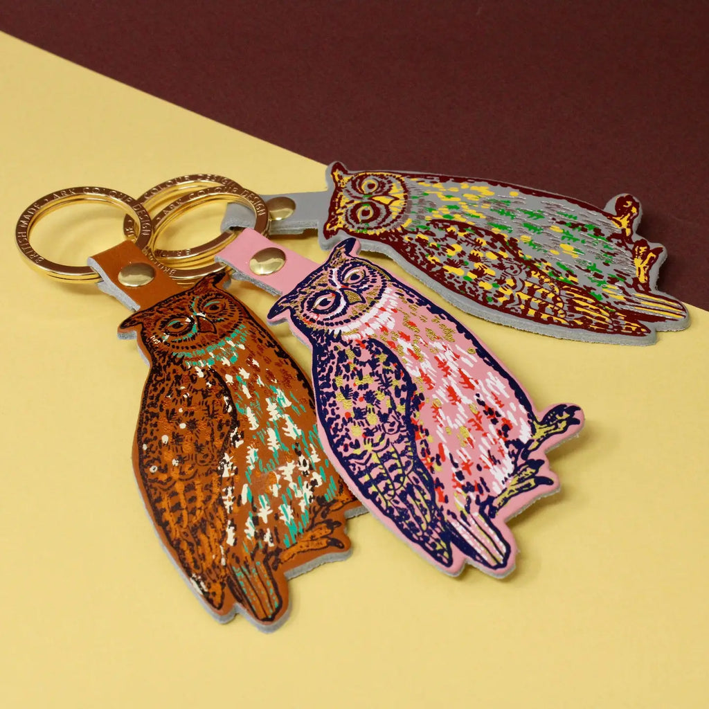 Ark Colour Design owl keychains in three color styles, shown with gold key rings on a yellow and brown background.