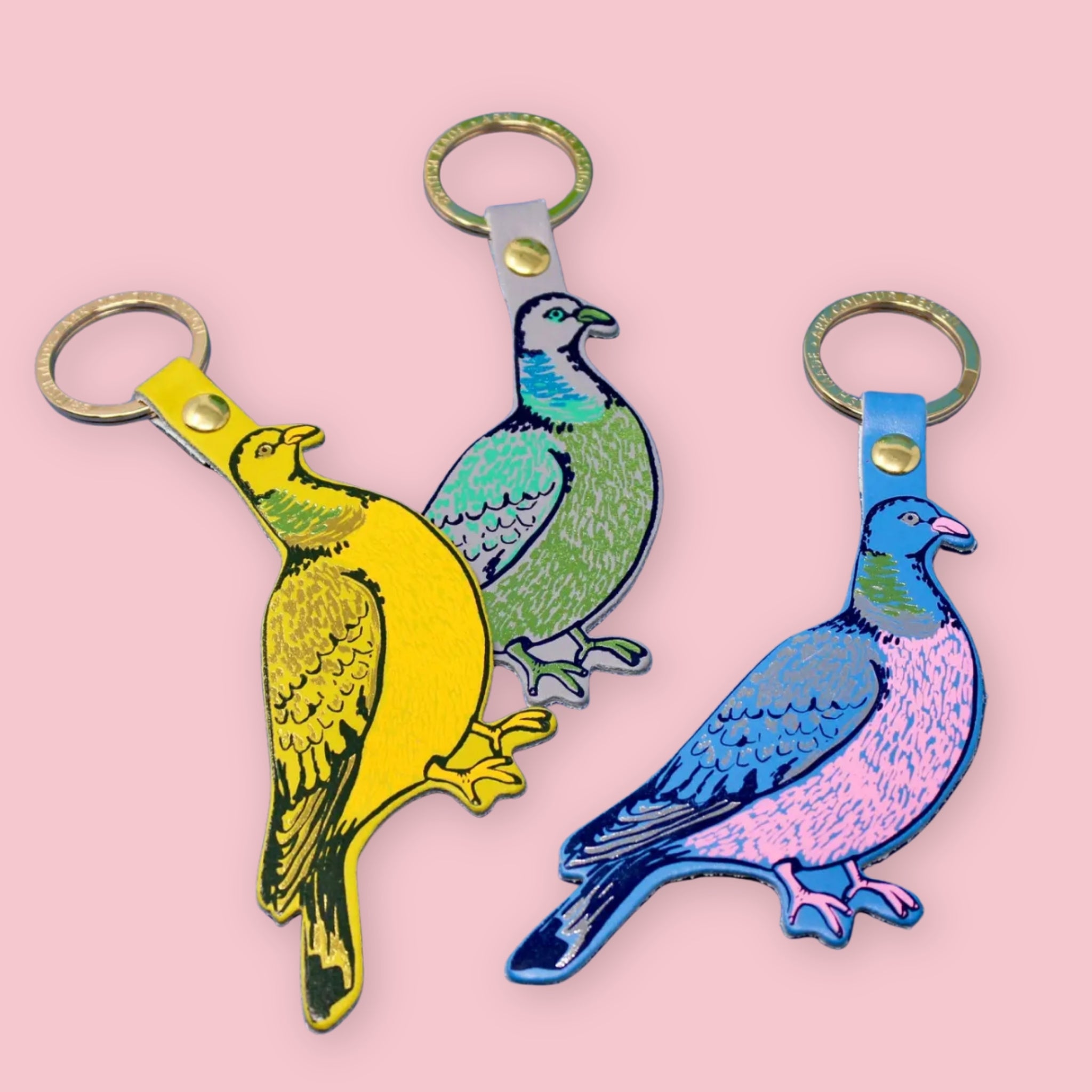 Three colorful pigeon-shaped keychains in yellow, green, and blue-pink, arranged on a solid pink background.