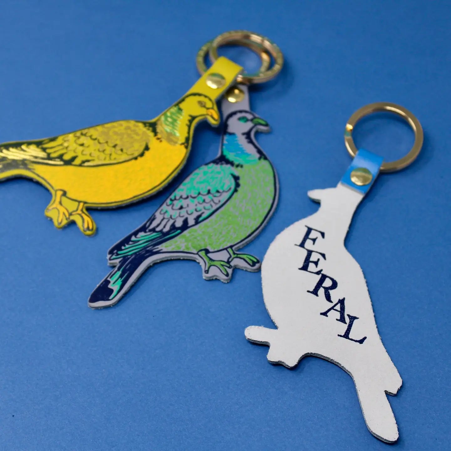Three bird-shaped keychains on a blue background with one showing its backside with the text reading "FERAL"