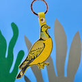 A yellow pigeon-shaped keychain with black outlines and a green iridescent neck patch, attached to a yellow loop with a metal ring, shown against a blue background with paper cutout seaweed shapes.