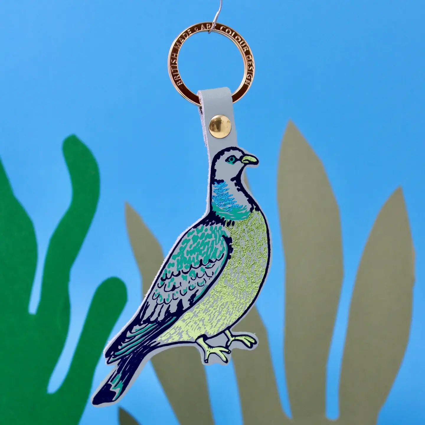 A bird-shaped keychain featuring a stylized pigeon with teal, green, and pale yellow coloring, attached to a white strap and a gold-tone ring. The keychain is photographed against a bright blue background with green and beige leaf silhouettes.