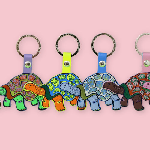Ark Colour Design tortoise keychains in assorted colors, lined up with gold key rings on a pink background.