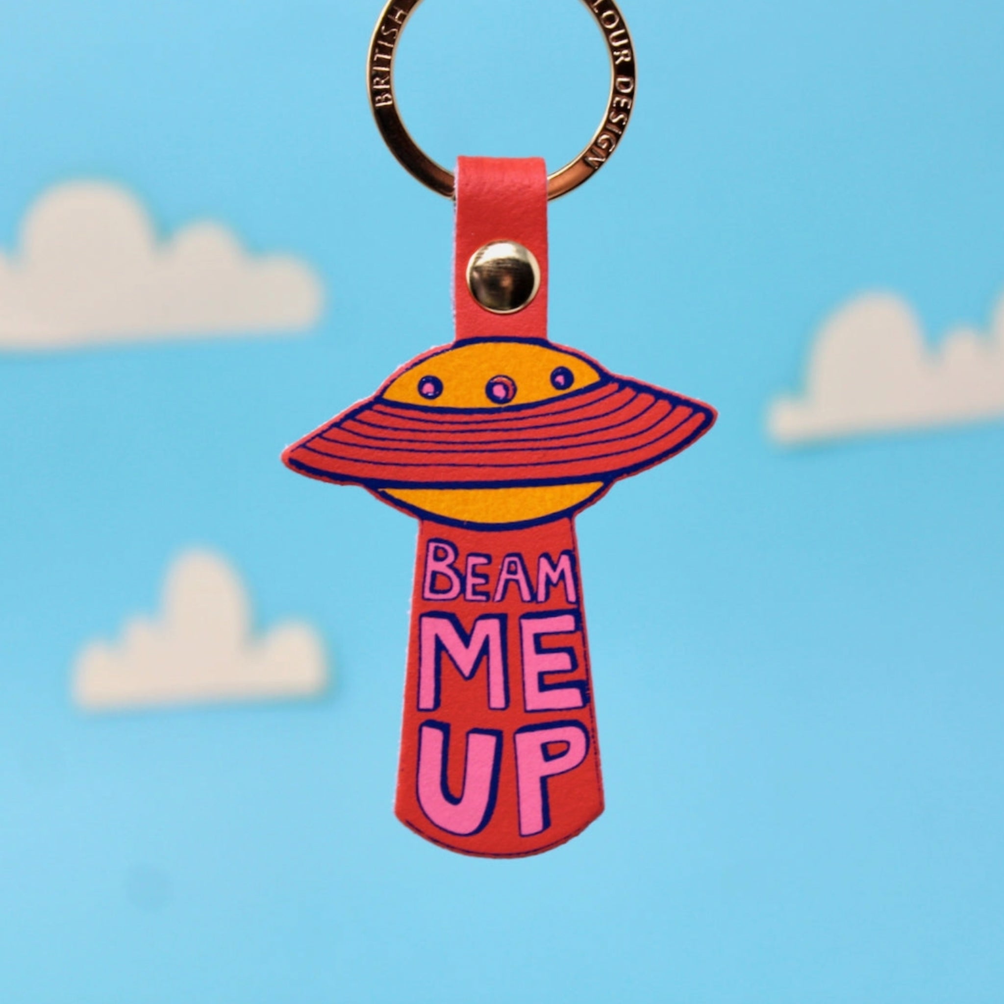 Ark Colour Design UFO keychain with coral strap, gold ring, and “Beam Me Up” text on a blue sky background.