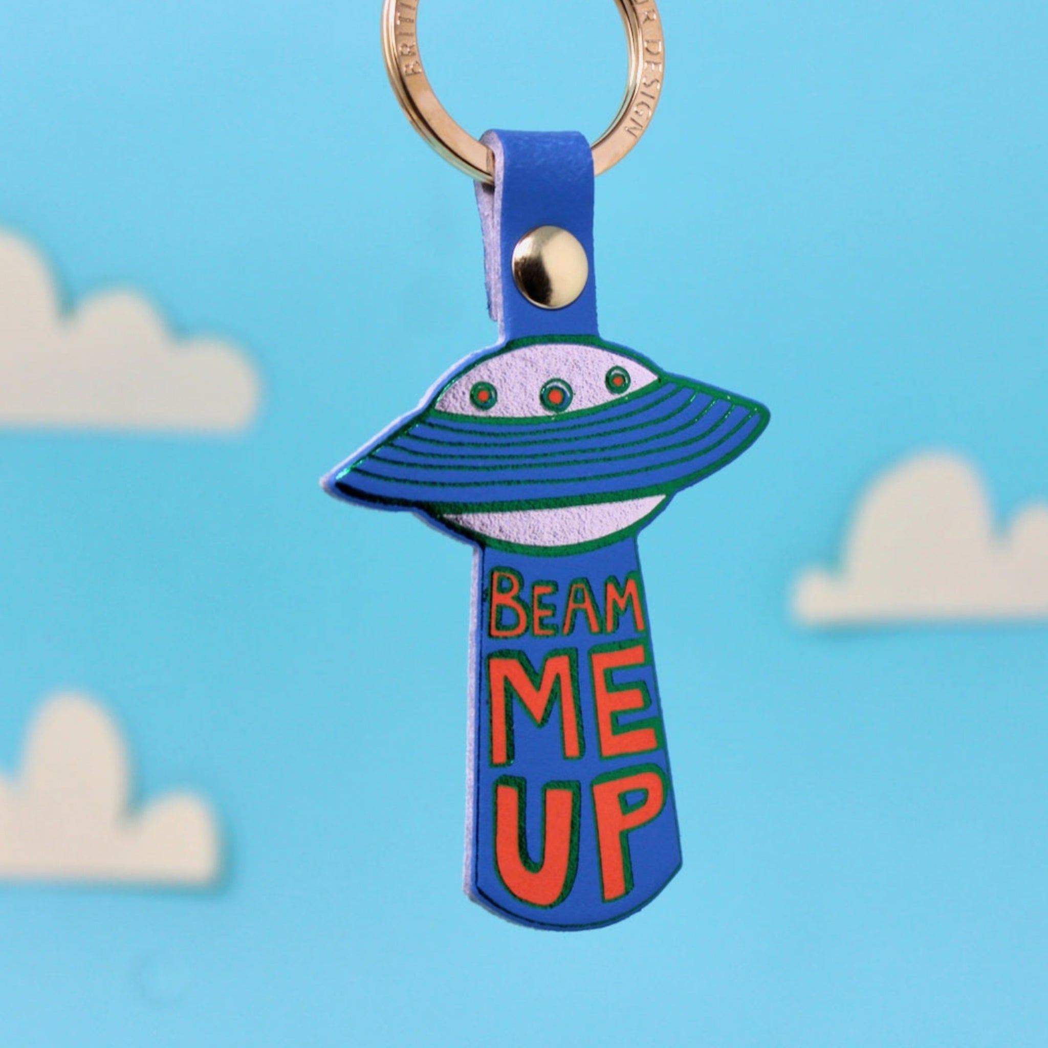 Ark Colour Design UFO keychain with blue strap, gold ring, and “Beam Me Up” text on a blue sky background.