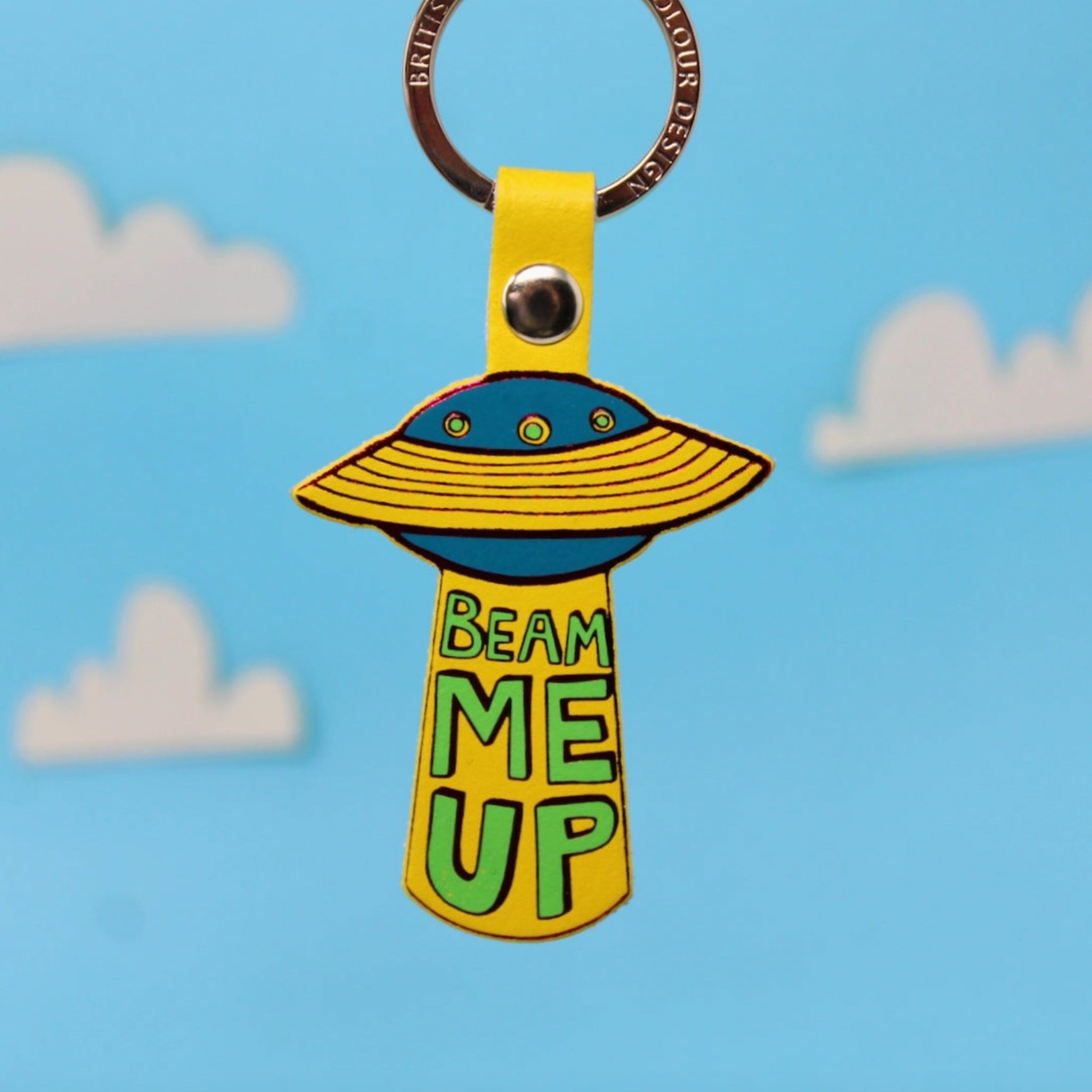 Ark Colour Design UFO keychain with yellow strap, gold ring, and “Beam Me Up” text on a blue sky background.