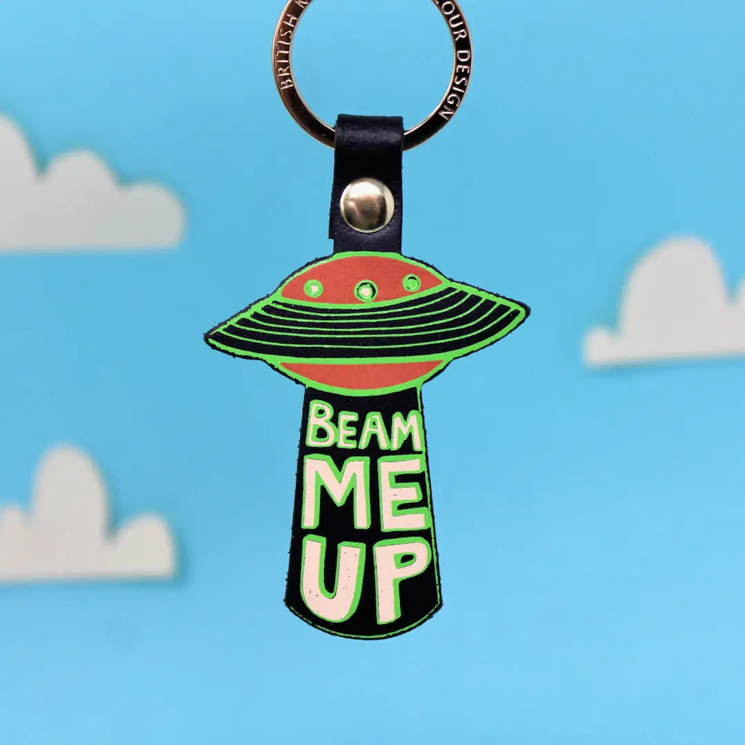 Ark Colour Design UFO keychain with black strap, gold ring, and “Beam Me Up” text on a blue sky background.