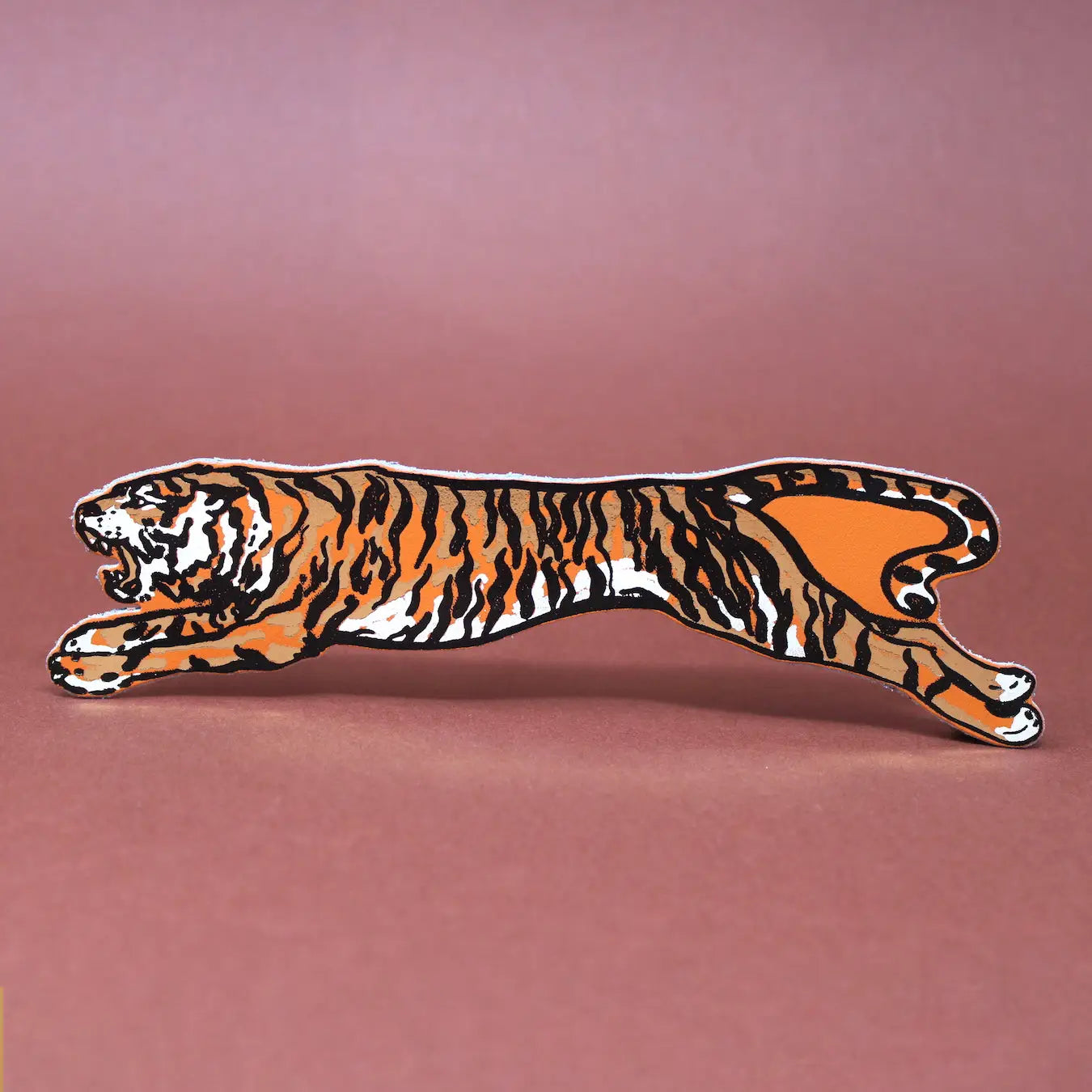 Leaping tiger-shaped leather bookmark with tan, black, and cream stripes, displayed horizontally on a warm brown background.