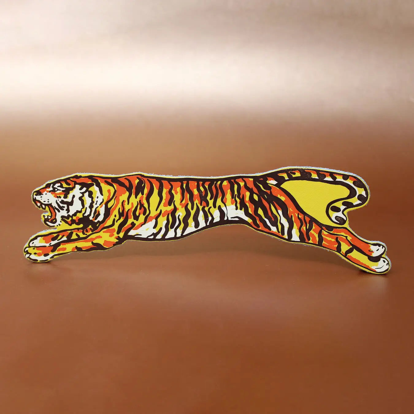 Leaping tiger-shaped leather bookmark with orange, black, and cream stripes, displayed horizontally on a warm brown background.