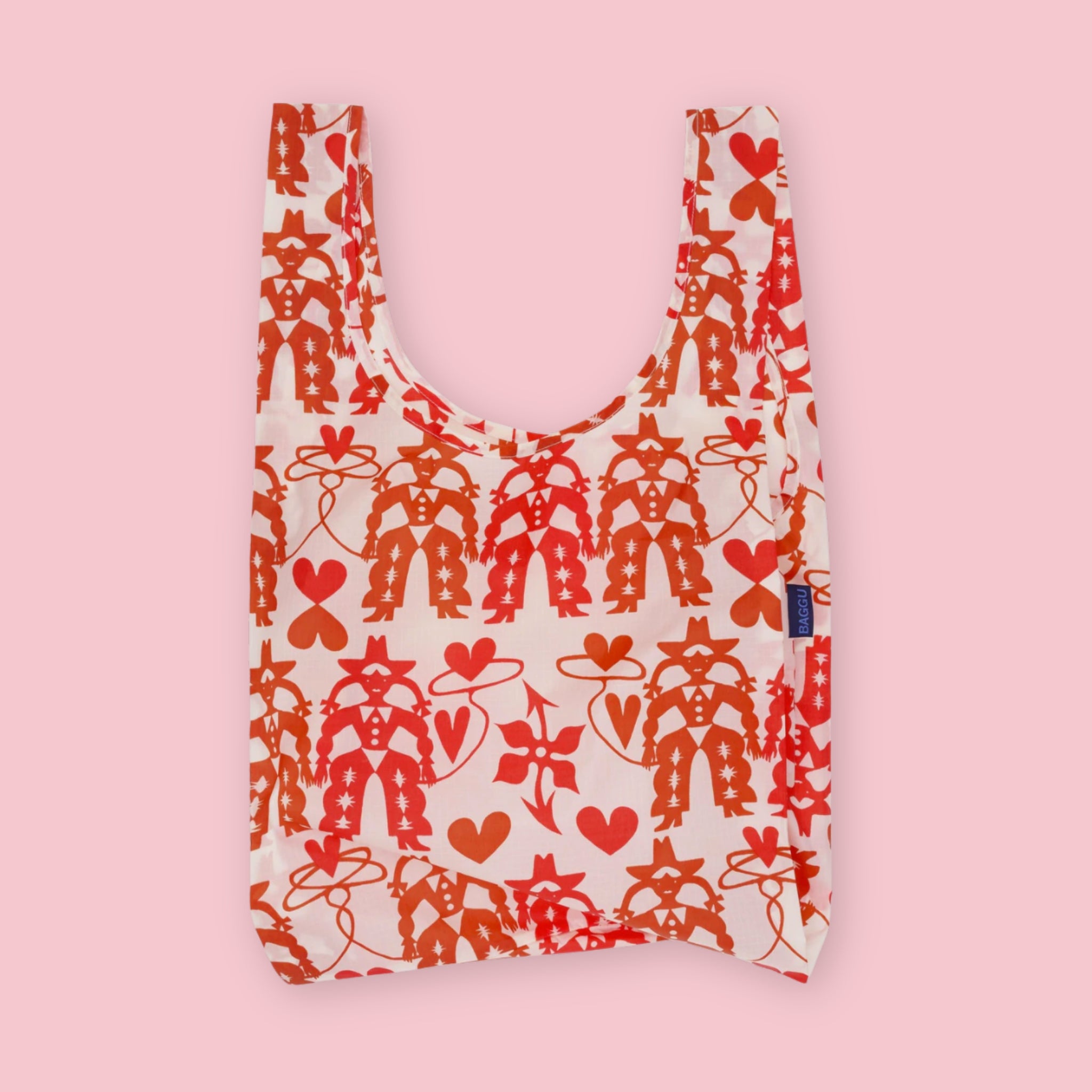 Baggu standard tote in the Cowgirl print, featuring a repeating pattern of cowgirl figures, hearts, and lasso motifs in red and orange on a cream background, on a pink background.