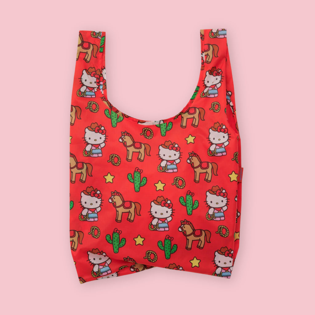 Baggu standard tote in the Hello Kitty Western print, featuring Hello Kitty in a cowgirl outfit with horses, cacti, lassos, and stars on a red background, on a pink background.