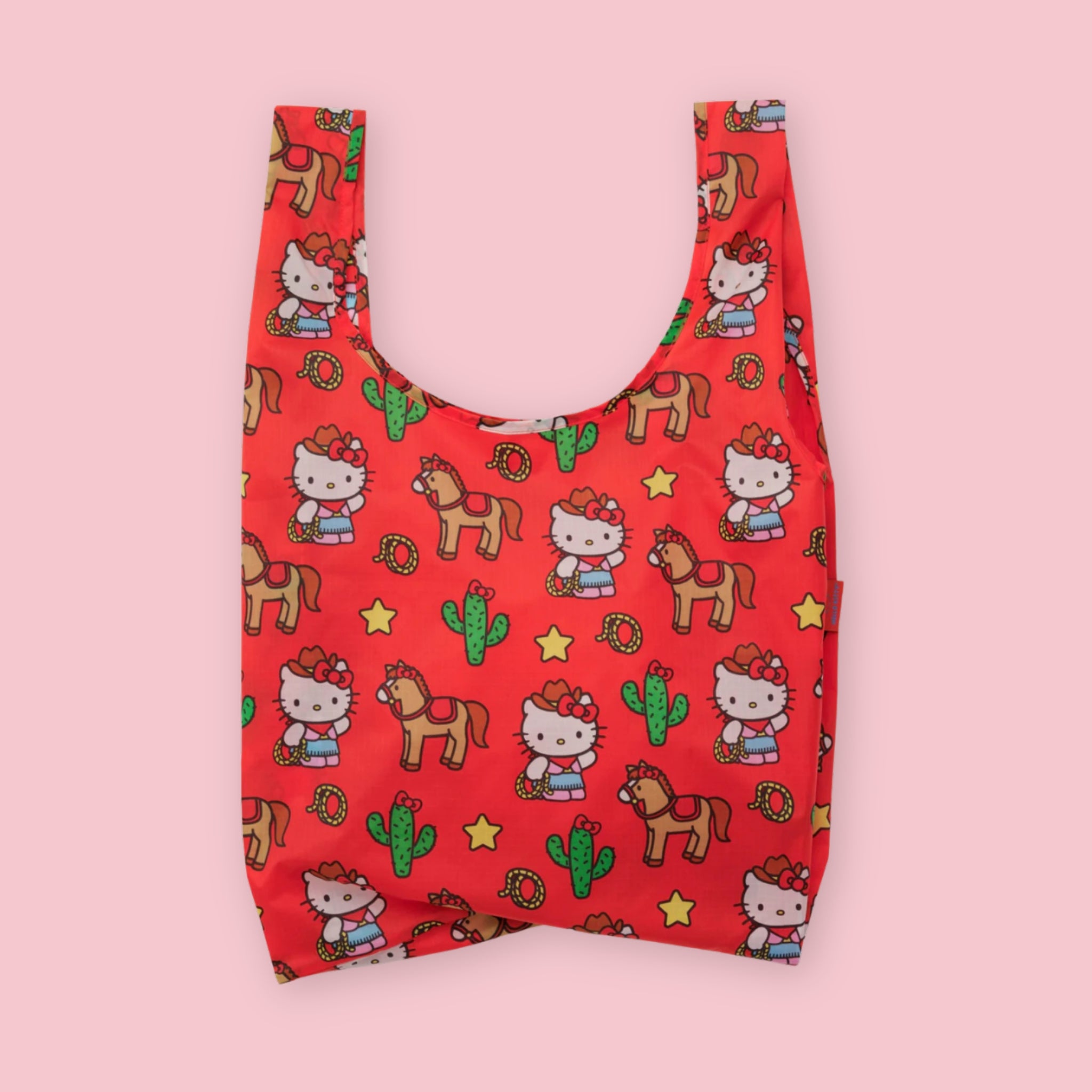 Baggu standard tote in the Hello Kitty Western print, featuring Hello Kitty in a cowgirl outfit with horses, cacti, lassos, and stars on a red background, on a pink background.