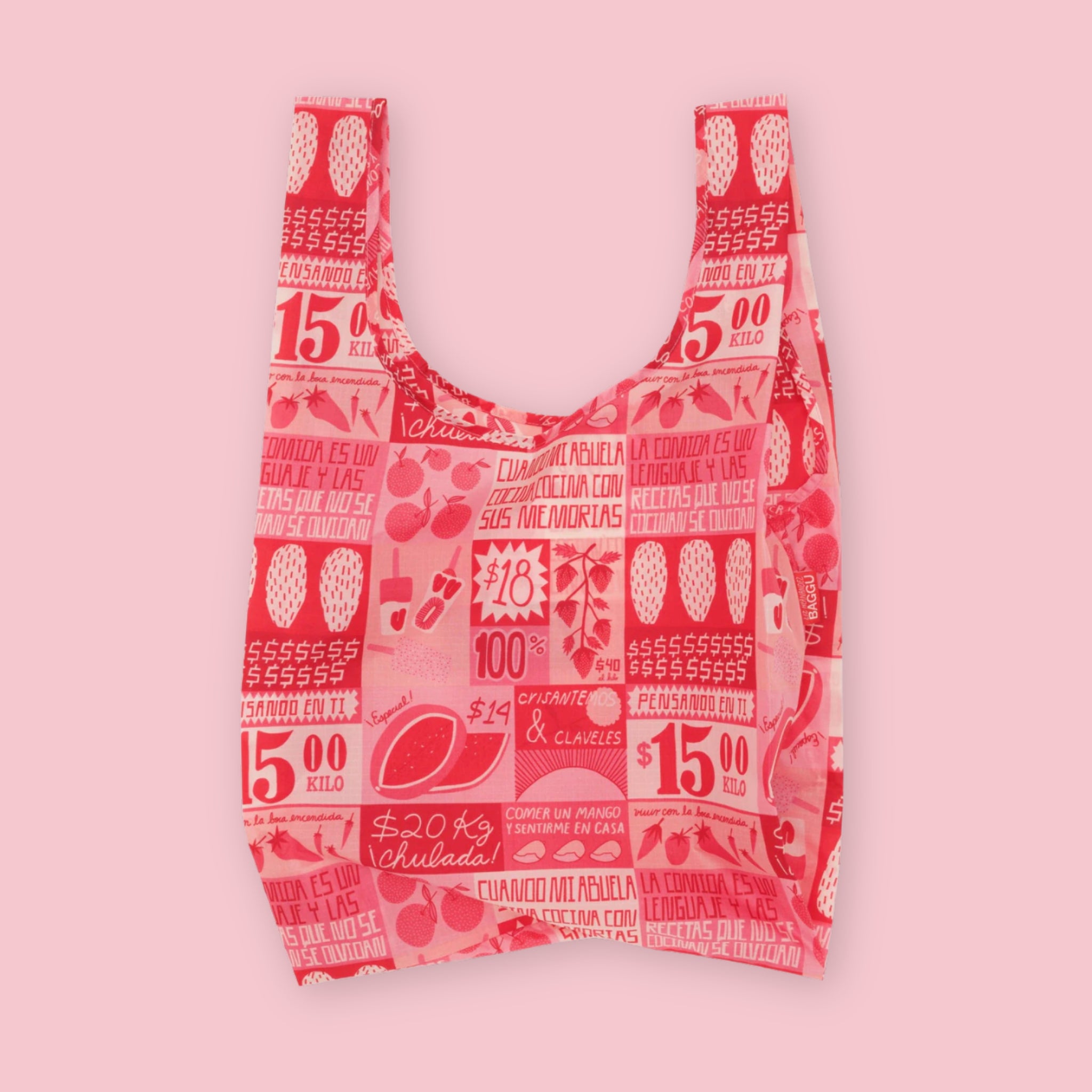 Baggu standard tote in the Mercado print, featuring a collage of market signage, price tags, fruit illustrations, and Spanish text phrases in red and pink on a cream background, on a pink background.