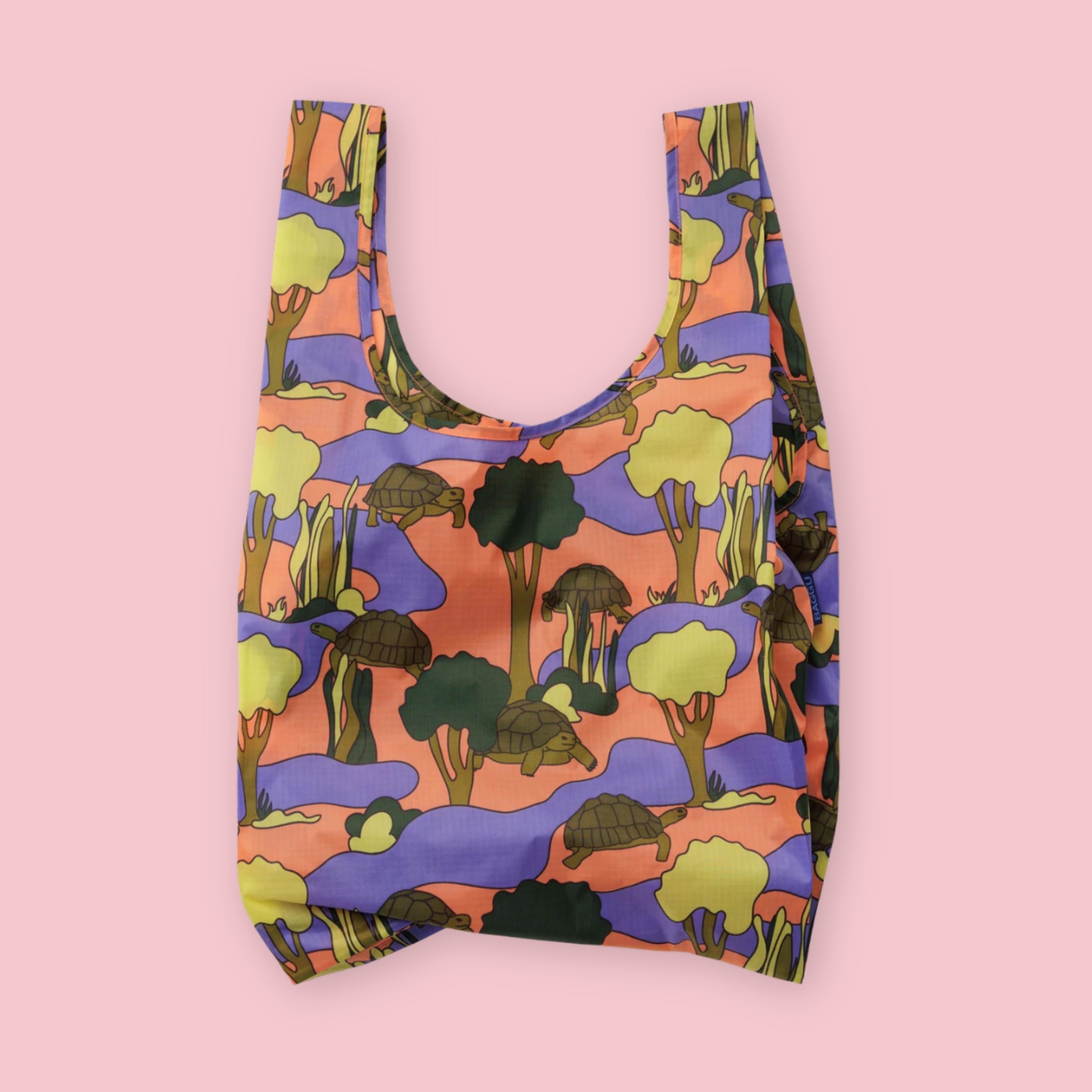 Baggu standard tote in the Turtle print, featuring a repeating landscape pattern of tortoises, cacti, and trees in orange, purple, and olive green, on a pink background.