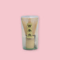 Bamboo matcha whisk stored in a clear plastic container with a natural wood whisk holder, displayed against a pastel pink background.