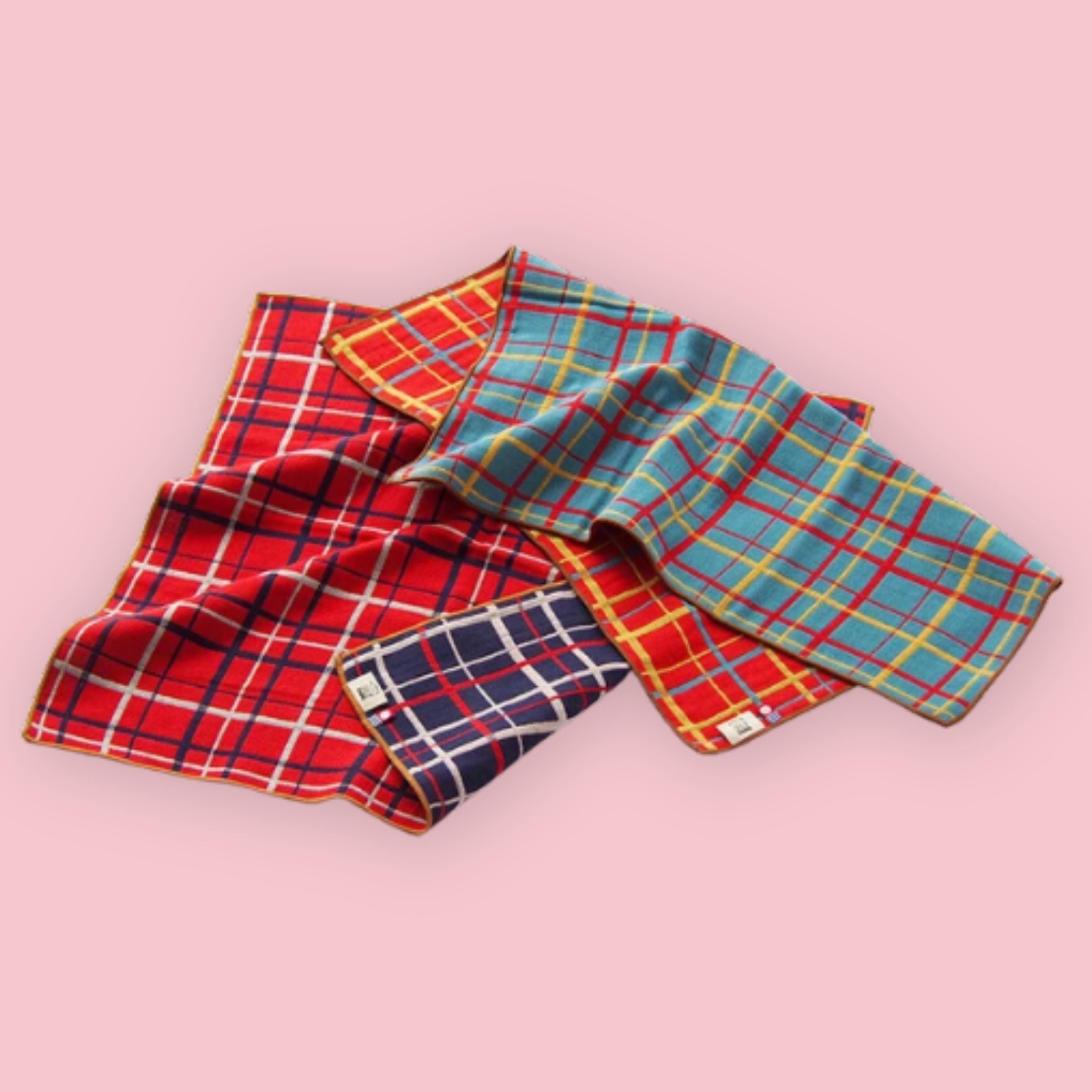 Imabari BLT Towels in Red Plaid and Blue Plaid styles loosely folded on a pink background