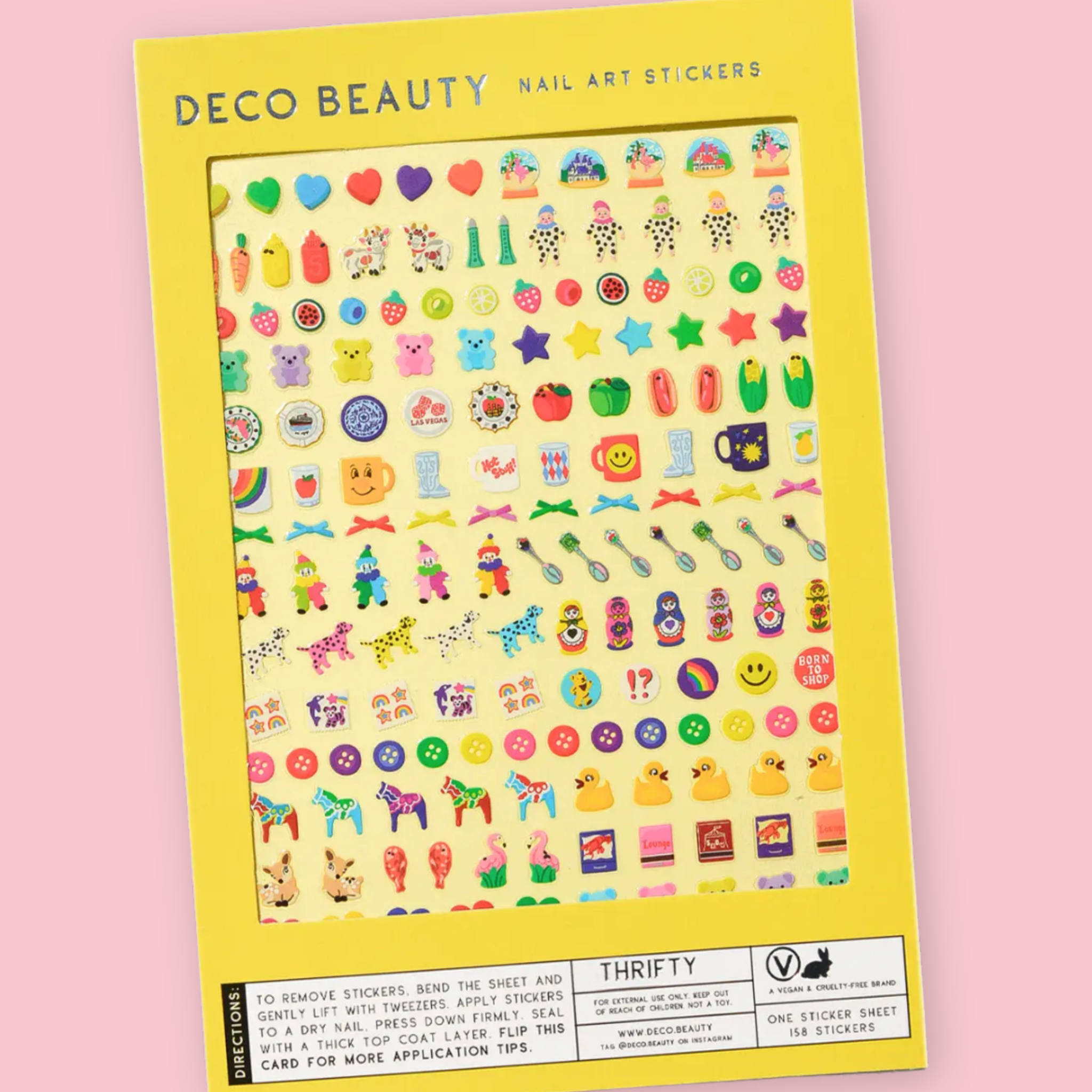 Deco Beauty Thrifty nail art sticker sheet packaging with 158 stickers depicting an eclectic mix of nostalgic motifs, hearts, snow globes, fruit, gummy bears, stars, mugs, matryoshka dolls, dalmatians, rubber ducks, flamingos, and buttons, on a yellow background.