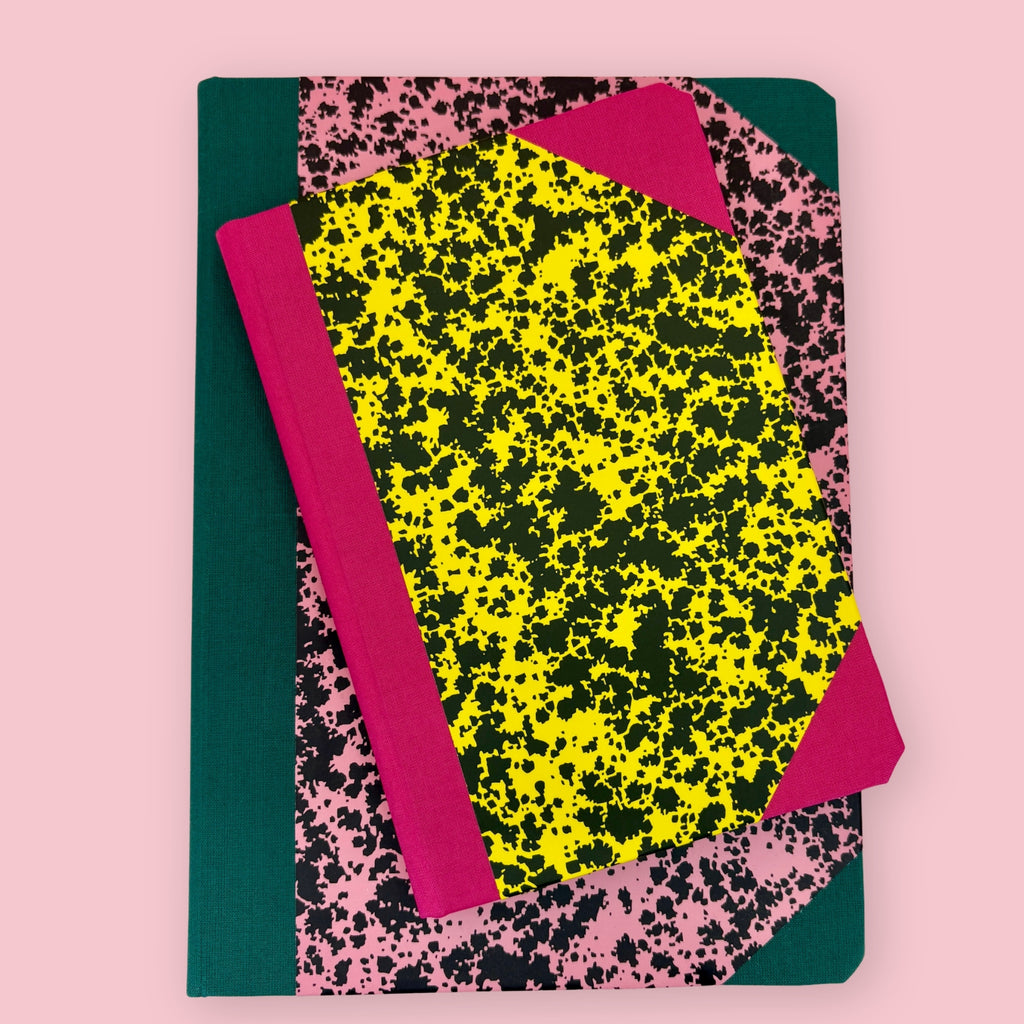 Two patterned notebooks with pink, yellow, black, and green covers, shown stacked against a pink background.