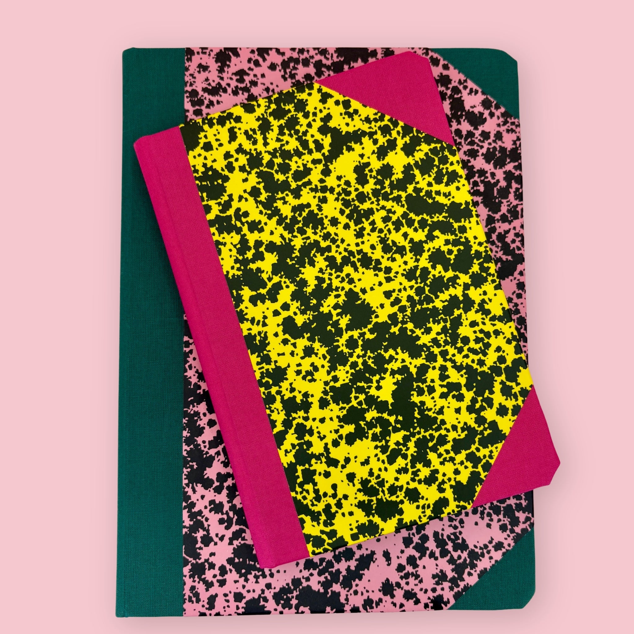 Two patterned notebooks with pink, yellow, black, and green covers, shown stacked against a pink background.