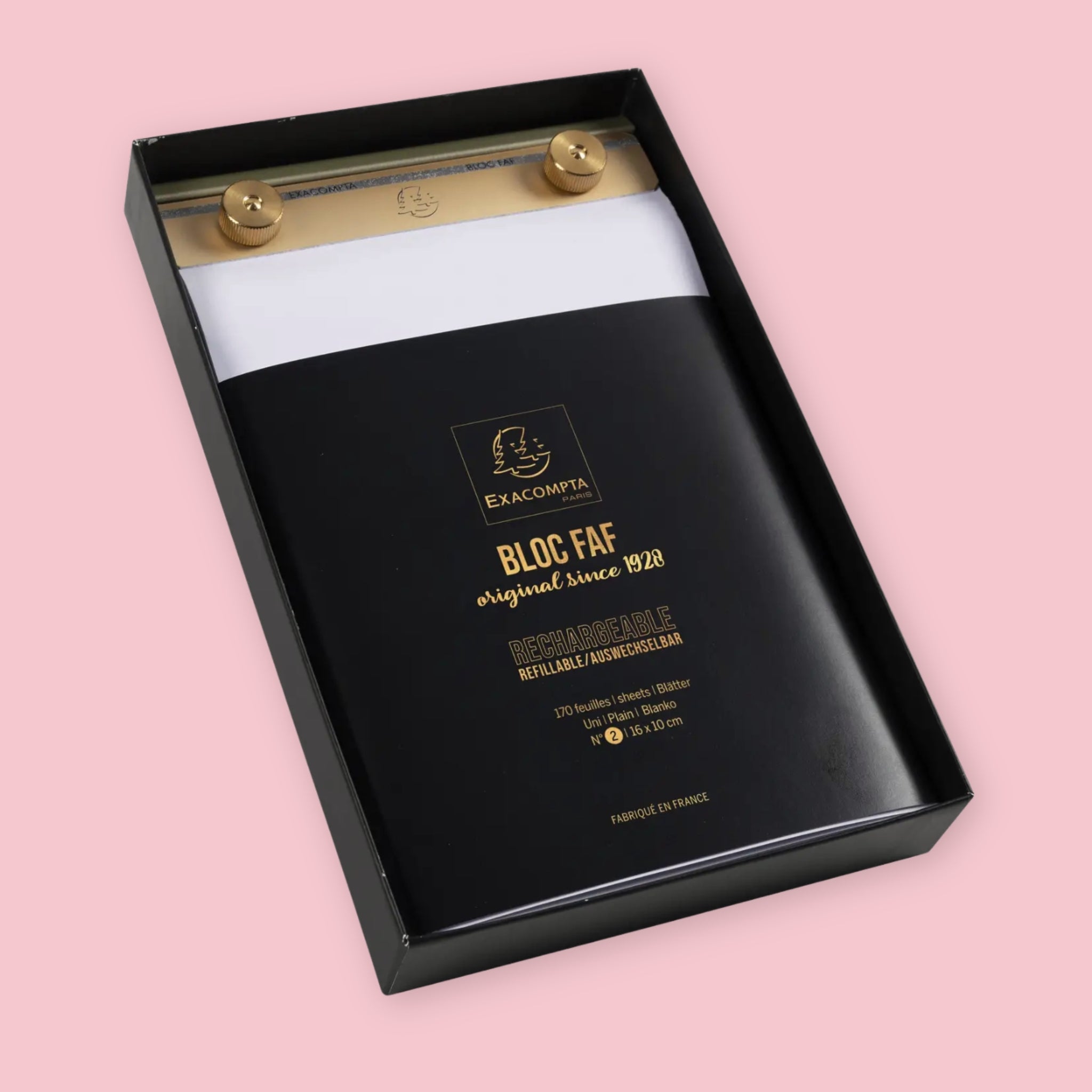 Exacompta Bloc FAF brass desk pad in its black and gold branded packaging box, showing the gold brass bar and knobs holding white blank paper, on a pink background.