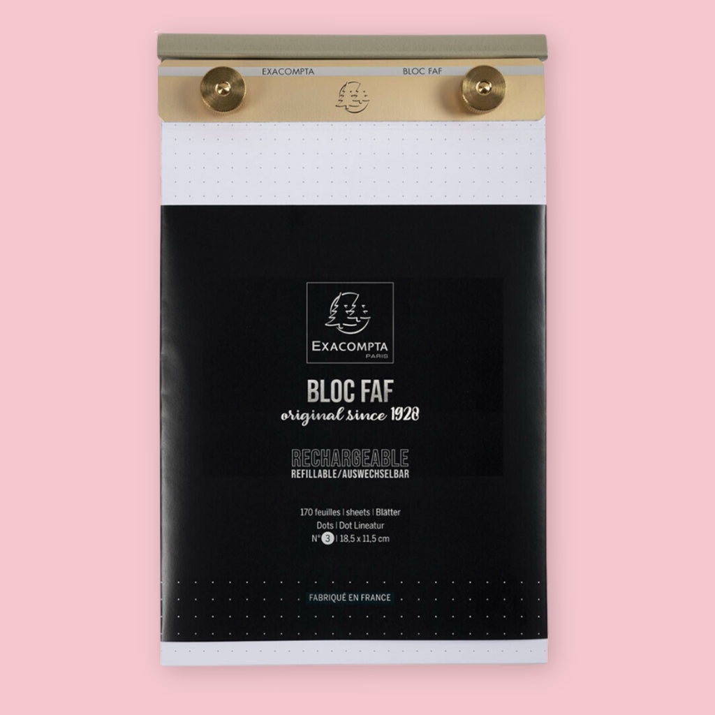 Exacompta Bloc FAF brass desk pad in the 5.25 x 8.25 dot grid size, showing the gold brass bar and knobs holding dot grid paper above the black branded packaging, on a pink background.