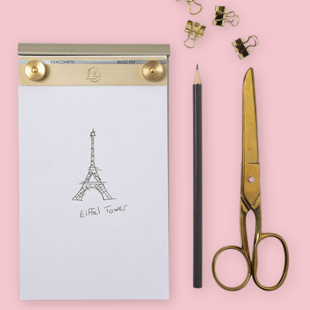 Exacompta brass desk pad with blank paper showing a hand-drawn Eiffel Tower sketch, styled with a pencil, gold scissors, and gold binder clips, on a pink background.