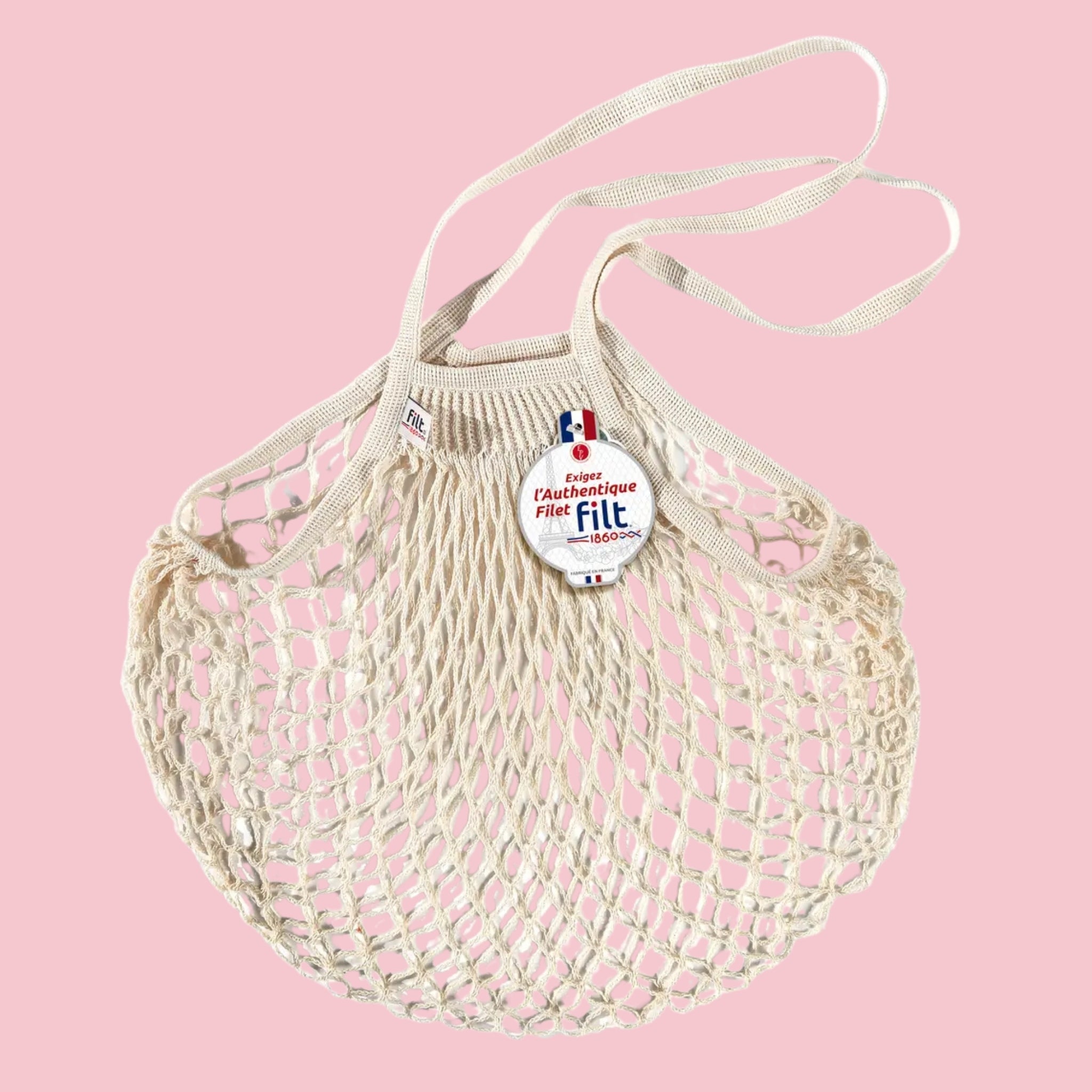 Filt French market mesh tote bag in natural with long handles and round brand tag, shown on a pink background.