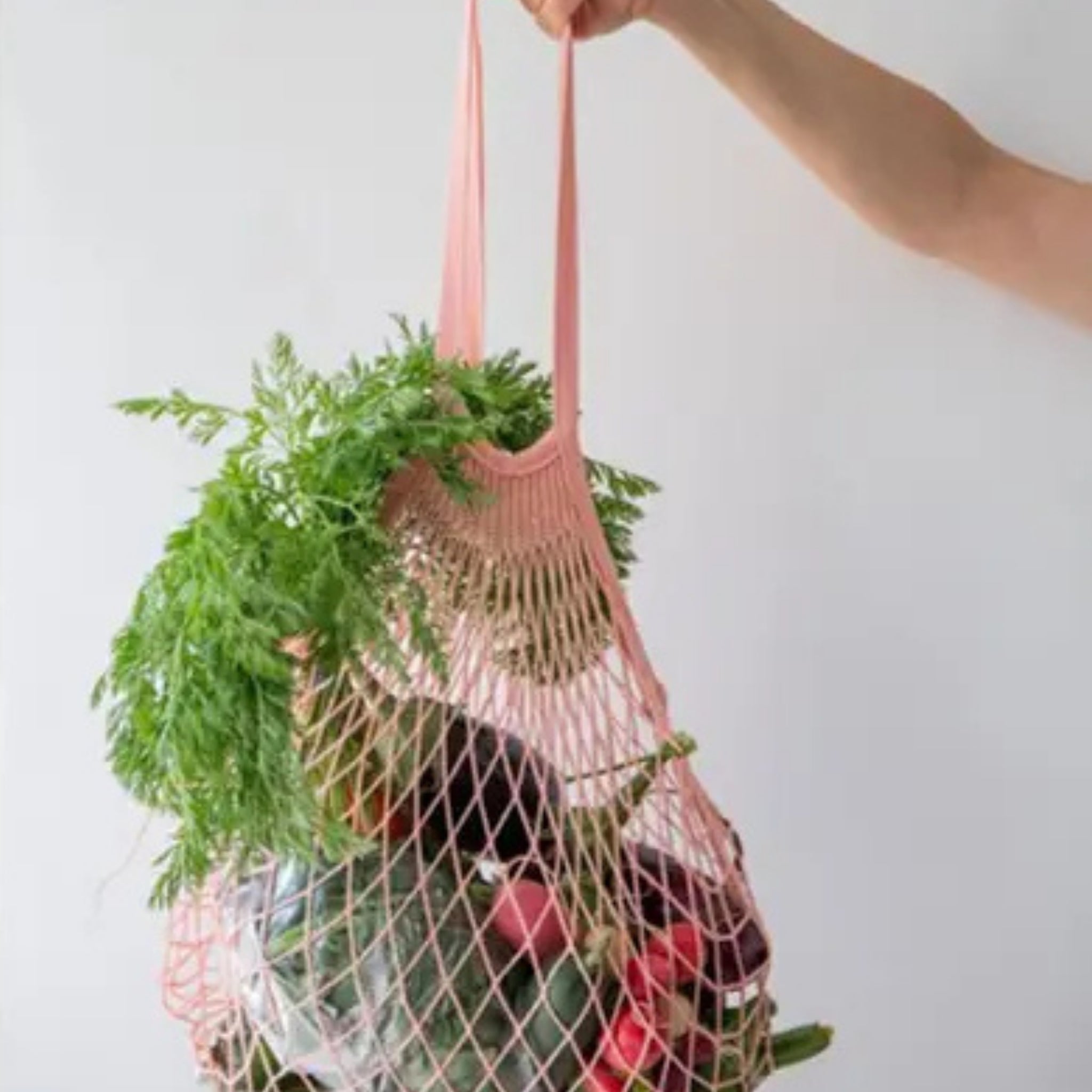 A pink mesh grocery bag being held by a hand, filled with green leafy vegetables and red radishes, against a plain white background.
