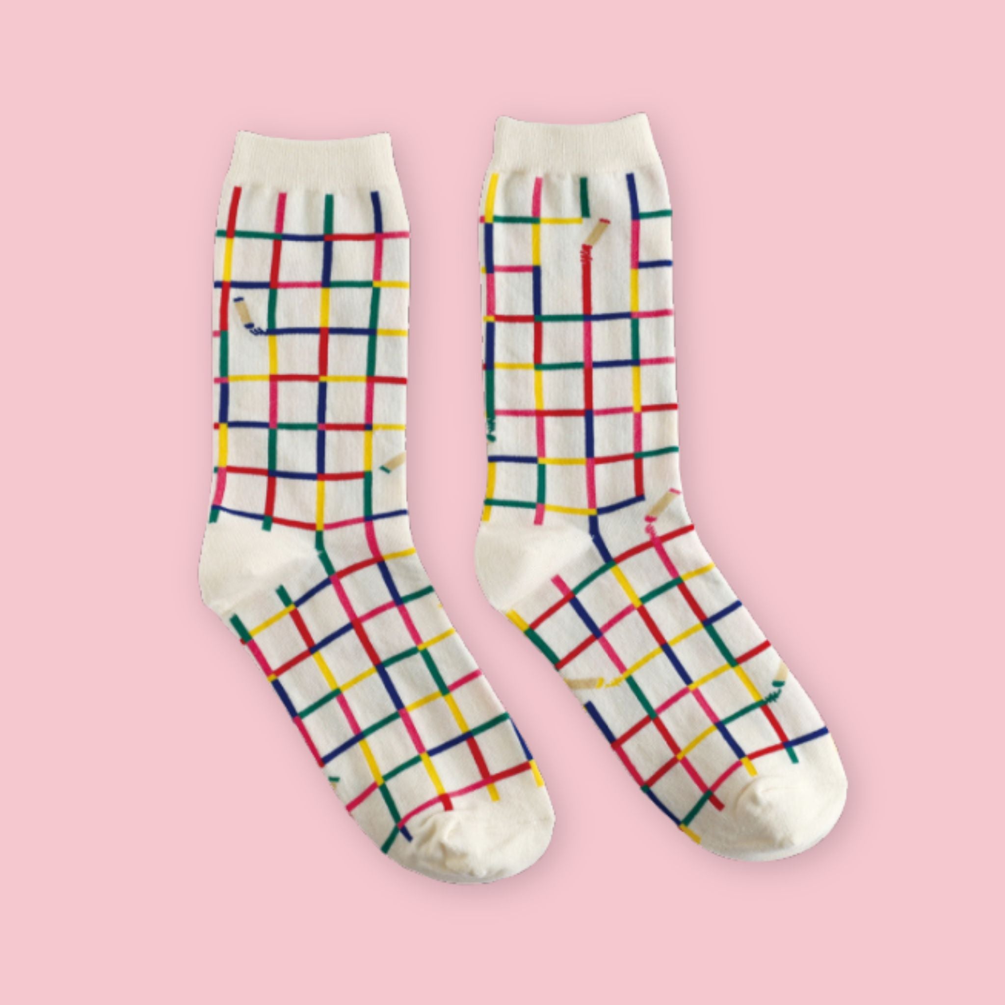 White crew socks with a colorful crayon-inspired grid pattern in red, blue, yellow, and green, displayed on a pastel pink background.