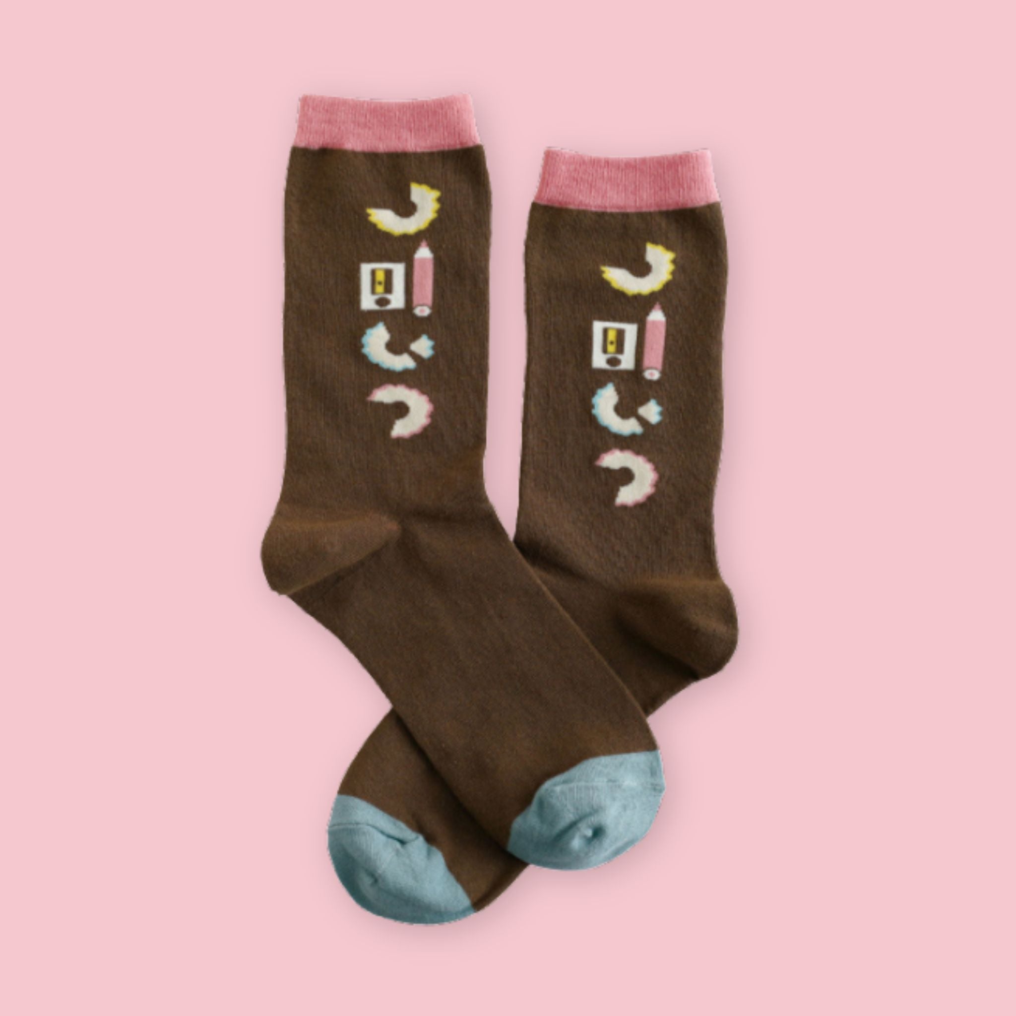Brown crew socks featuring a pencil sharpener, pink pencil, and curled pencil shavings design, with pink cuffs and light blue toes, displayed on a pastel pink background.