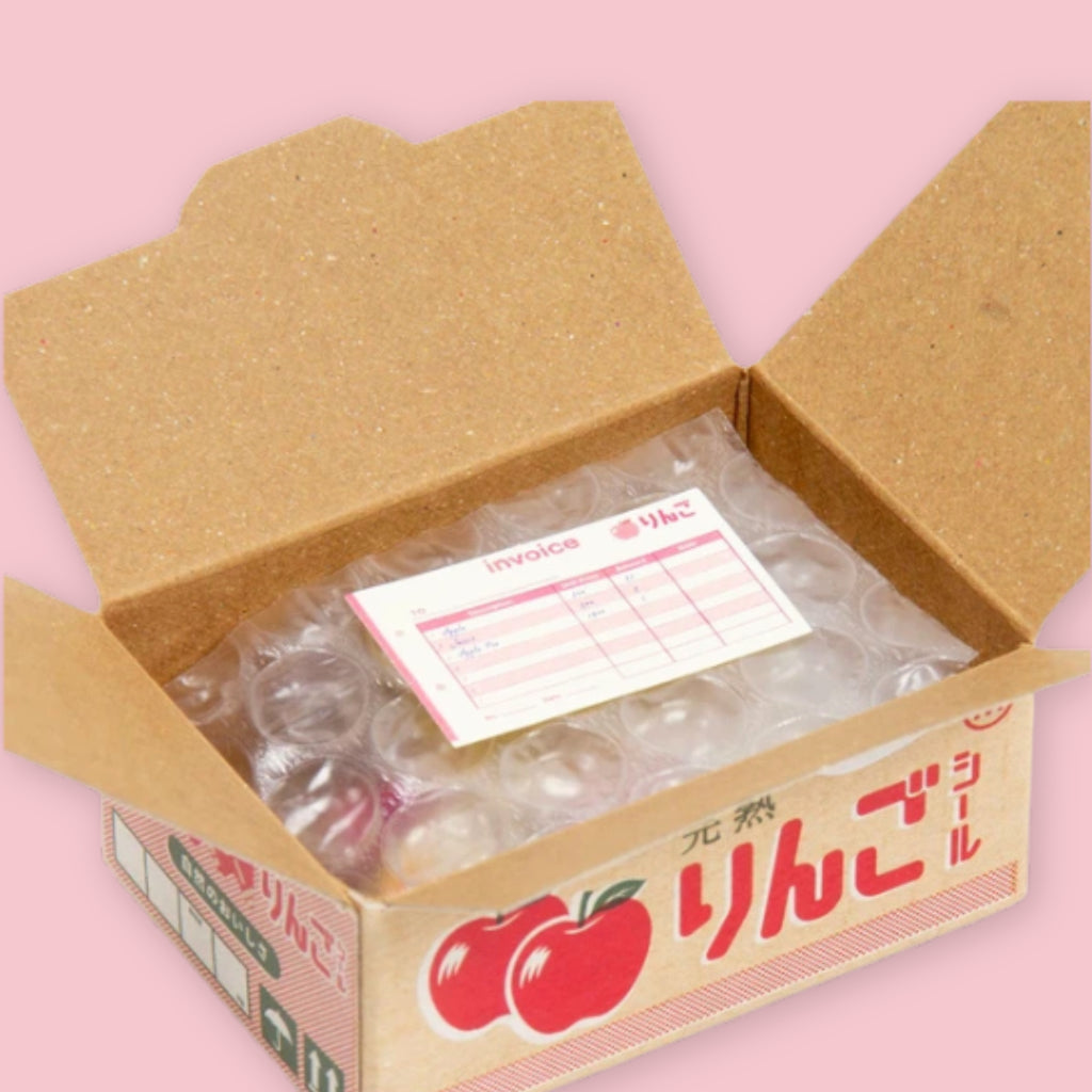 Open Hako sticker box showing bubble-wrapped stickers and a pink invoice inside, with the kraft brown Japanese apple "ringo" packaging visible on the exterior, on a pink background.