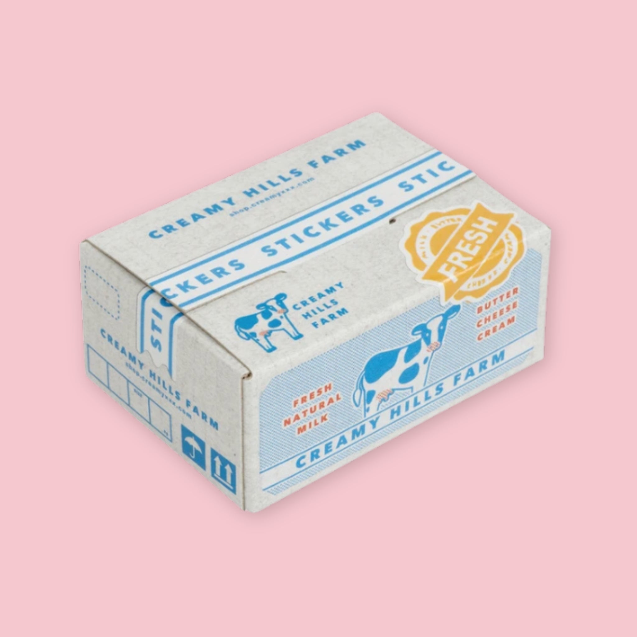 Box of Hako stickers designed to resemble vintage dairy packaging, with a white and blue color scheme, cow illustrations, "Creamy Hills Farm — Fresh Natural Milk, Butter, Cheese, Cream" branding, and a gold "Fresh" seal, on a pink background.