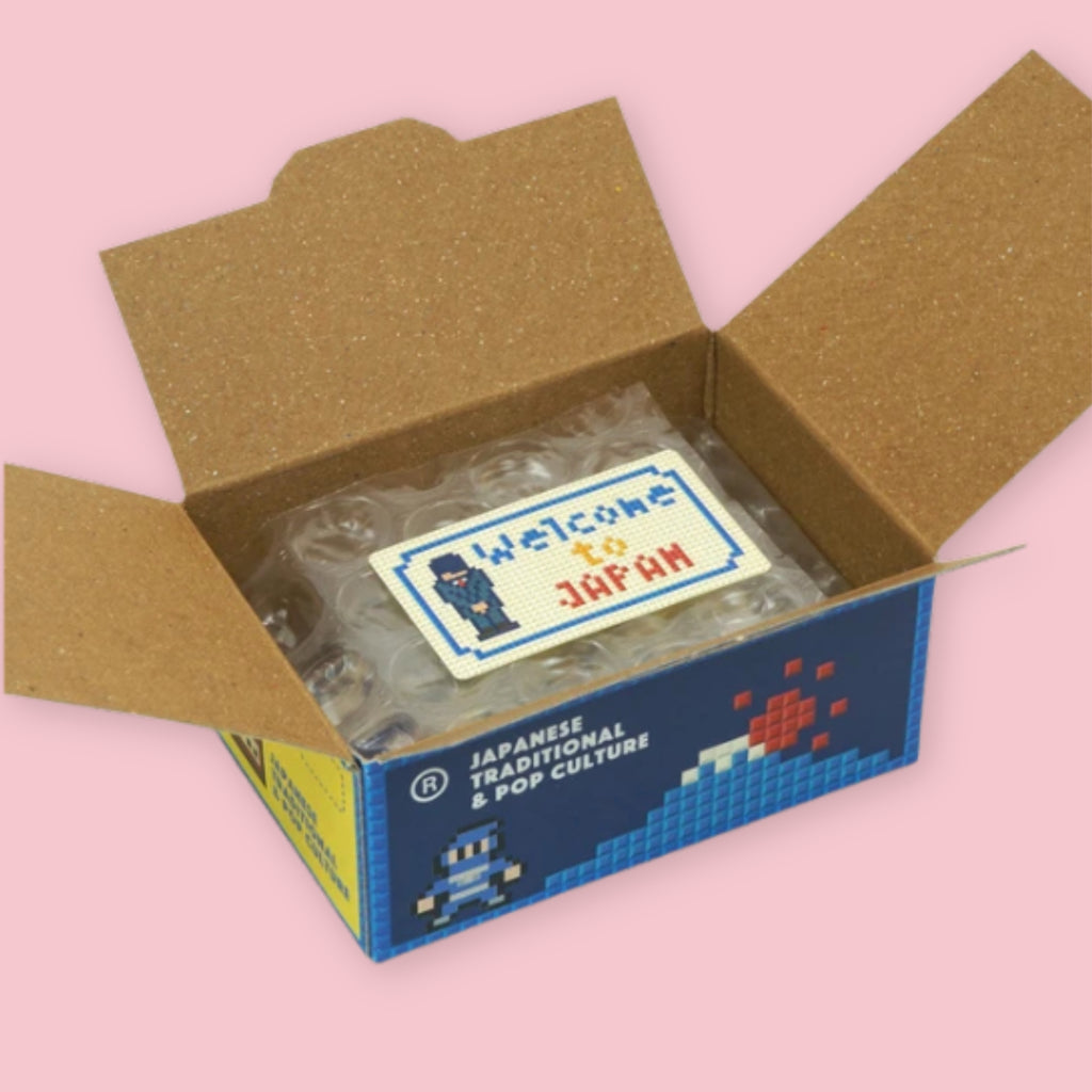 Open Hako sticker box showing bubble-wrapped stickers and a pixel art "Welcome to Japan" sticker on top, with the navy blue Japan Stickers Culture packaging visible on the exterior, on a pink background.