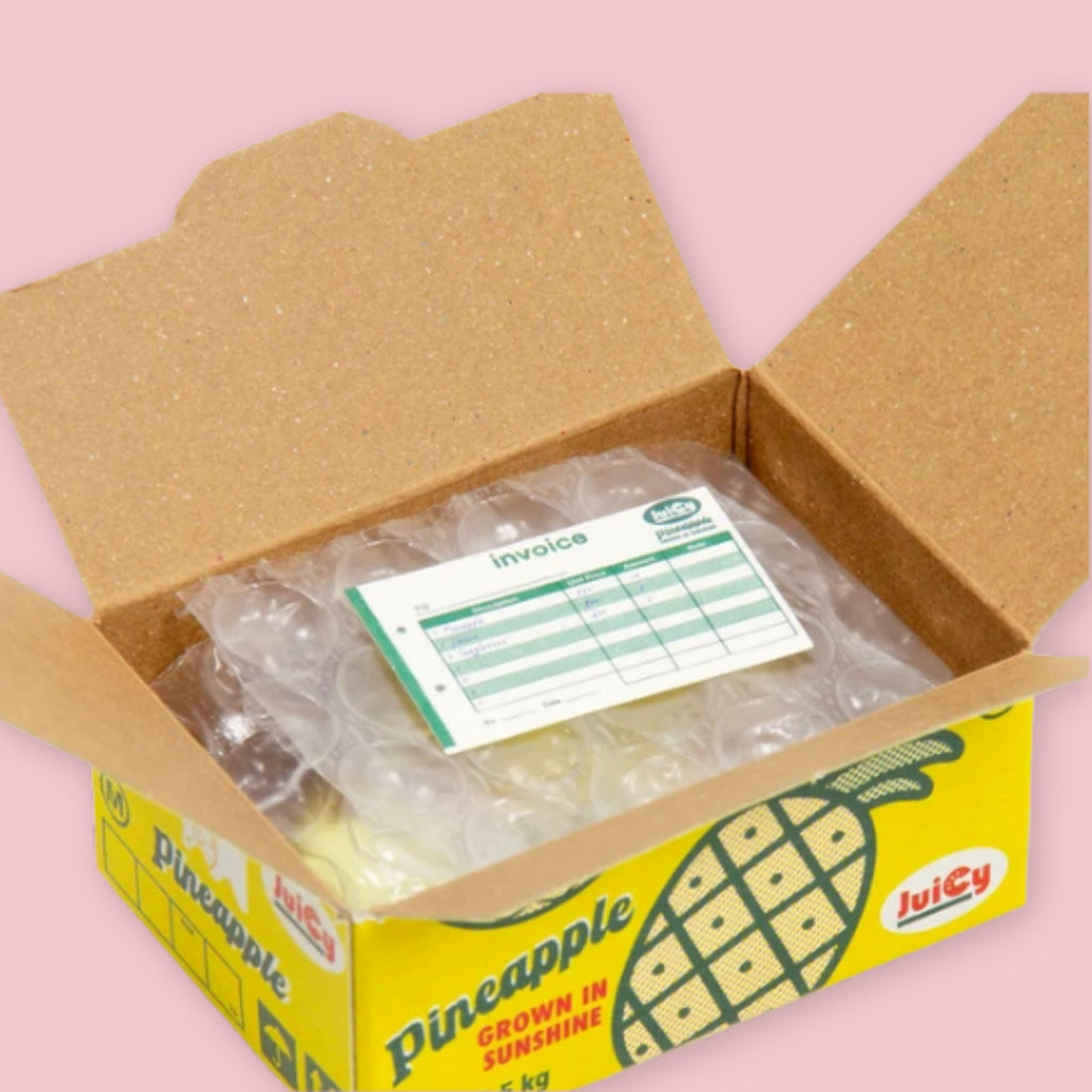 Open Hako sticker box showing bubble-wrapped stickers and an invoice inside, with the yellow pineapple "Juicy" packaging visible on the exterior, on a pink background.