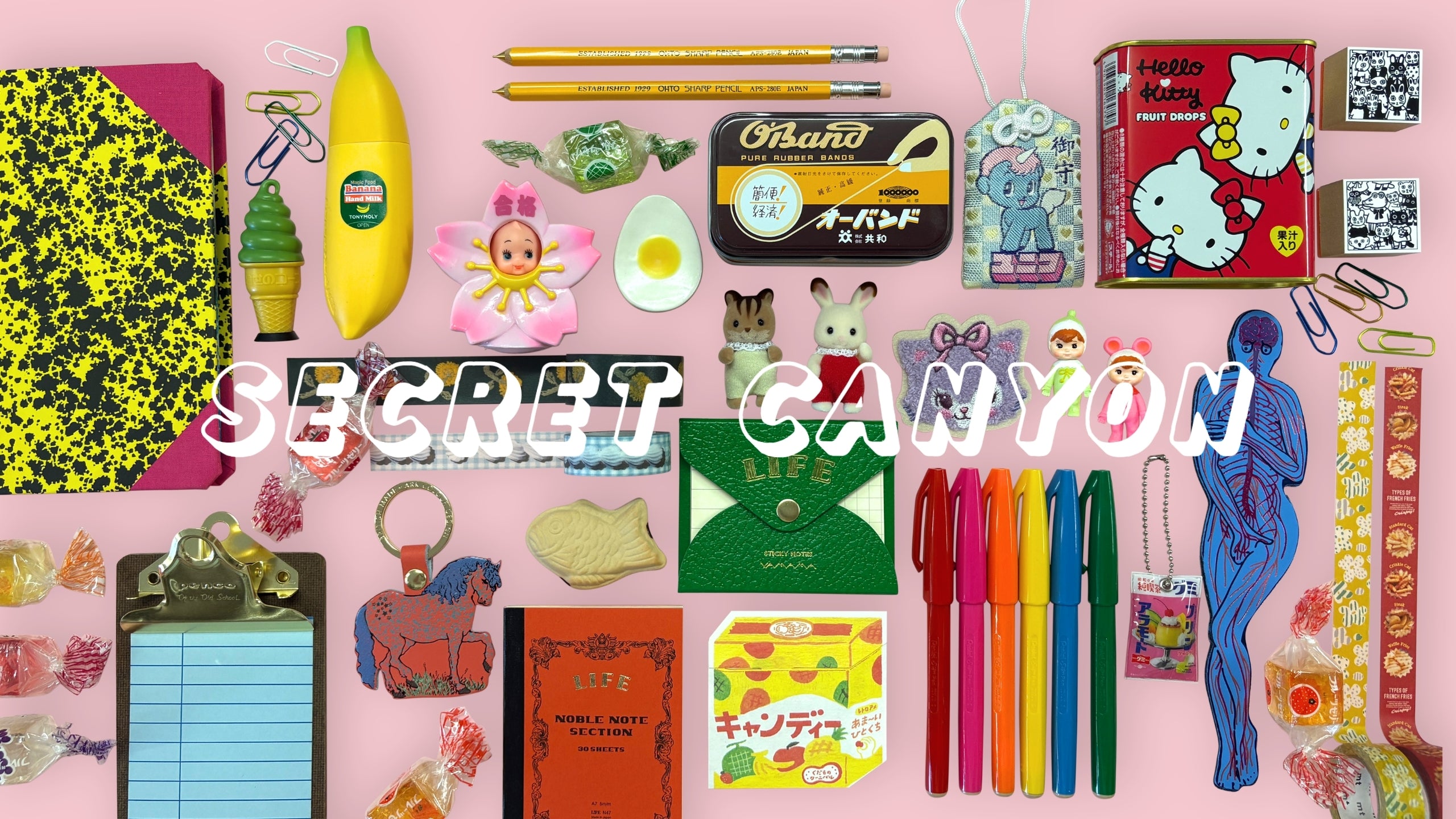 A flat lay of Japanese and European stationery and novelty goods spread across a pink background with "SECRET CANYON" overlaid in large white block letters, featuring items including a Hello Kitty Fruit Drops tin, TonyMoly banana lip balm, Sylvanian Families bunny figures, a Japanese O-Band rubber band tin, colorful felt-tip pens, a Life Noble Note notebook, a taiyaki-shaped eraser, a blue anatomical figure charm, wrapped candies, paper clips, yellow pencils, washi tape, and a red horse keychain