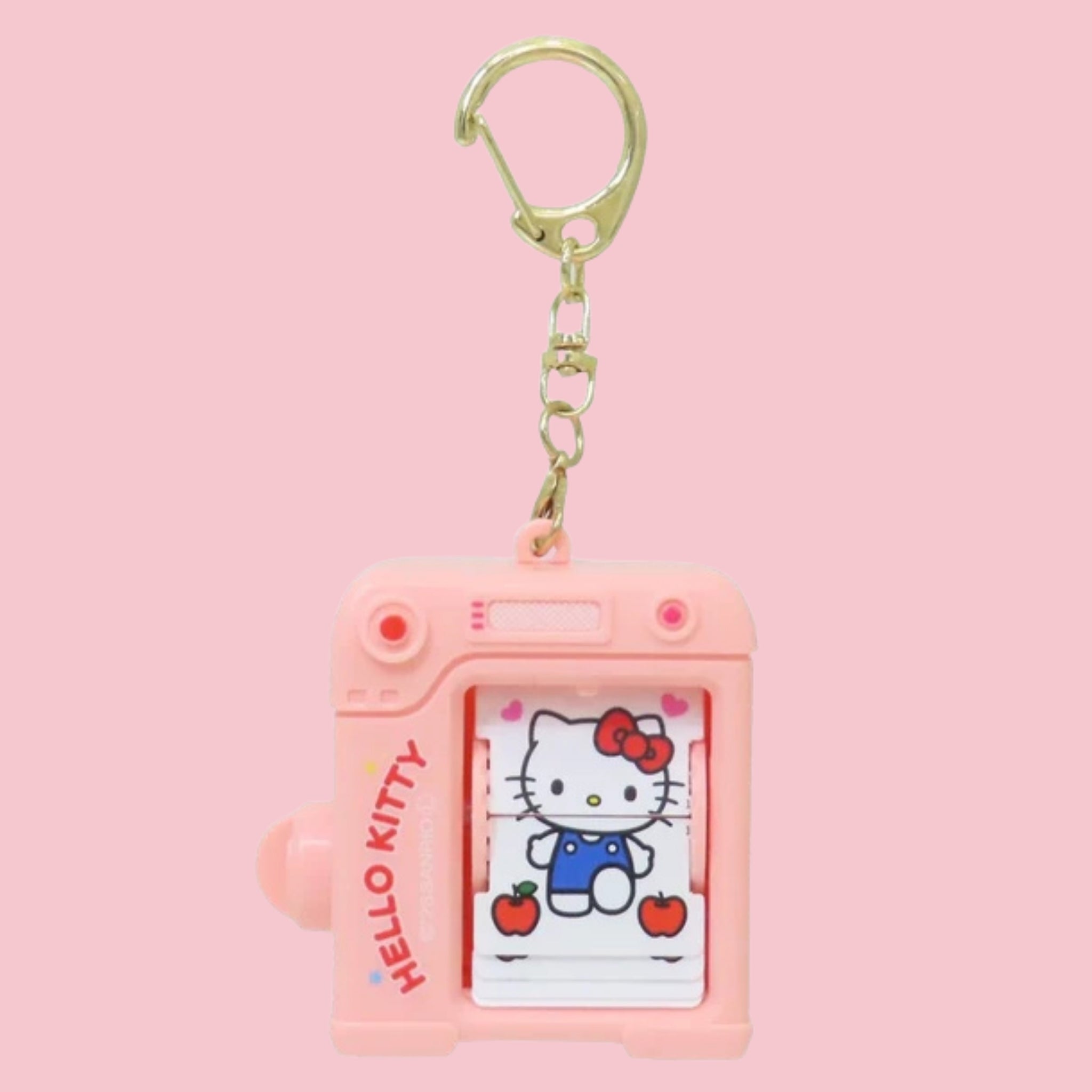 Pink plastic camera shaped keychain with a gold swivel clasp, showing Hello Kitty in blue overalls surrounded by hearts and apples on the flip book pages visible through the front window, against a solid pink background.