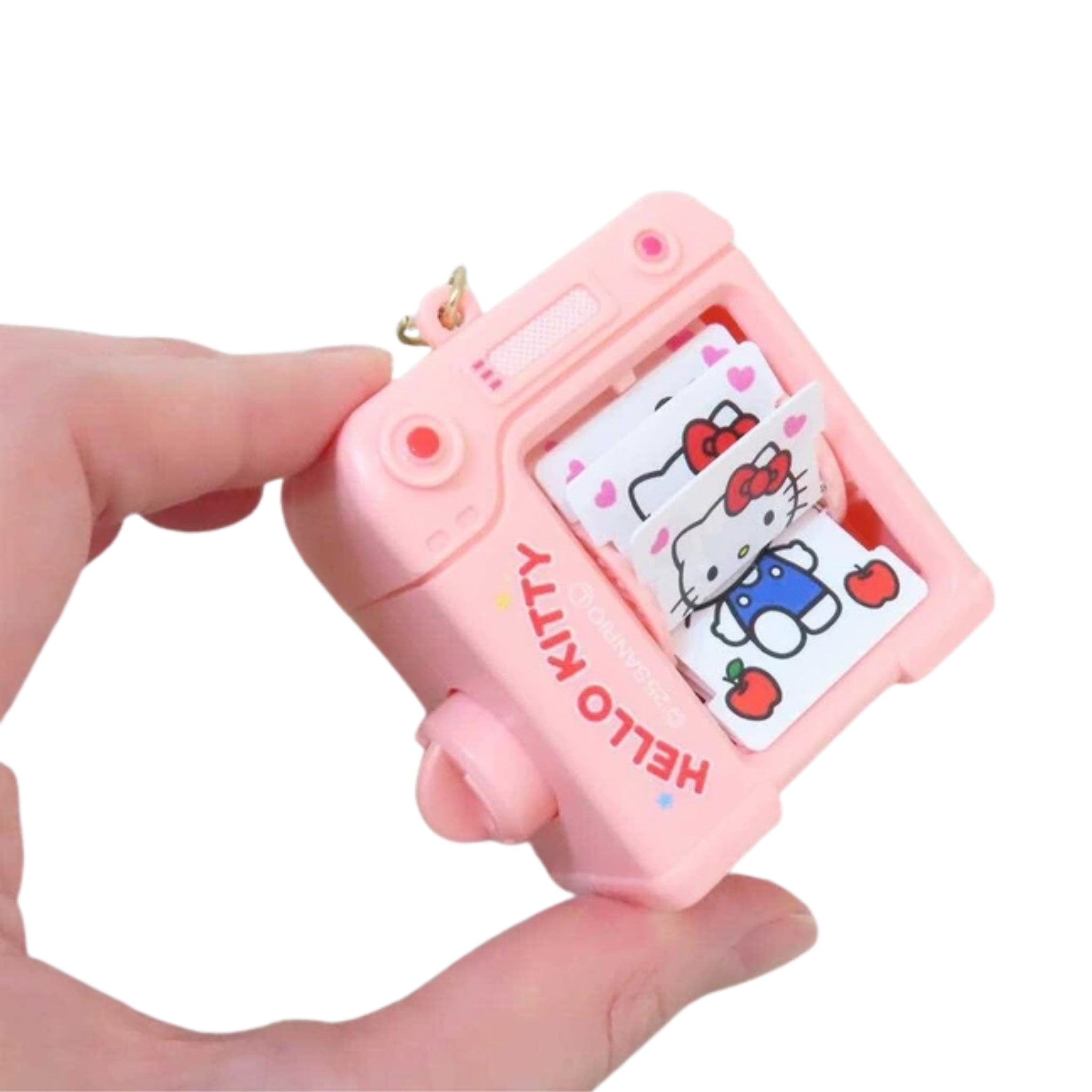 A hand holds the pink camera shaped keychain sideways, fanning out the small illustrated pages inside to show Hello Kitty in blue overalls surrounded by hearts and apples, against a plain white background.